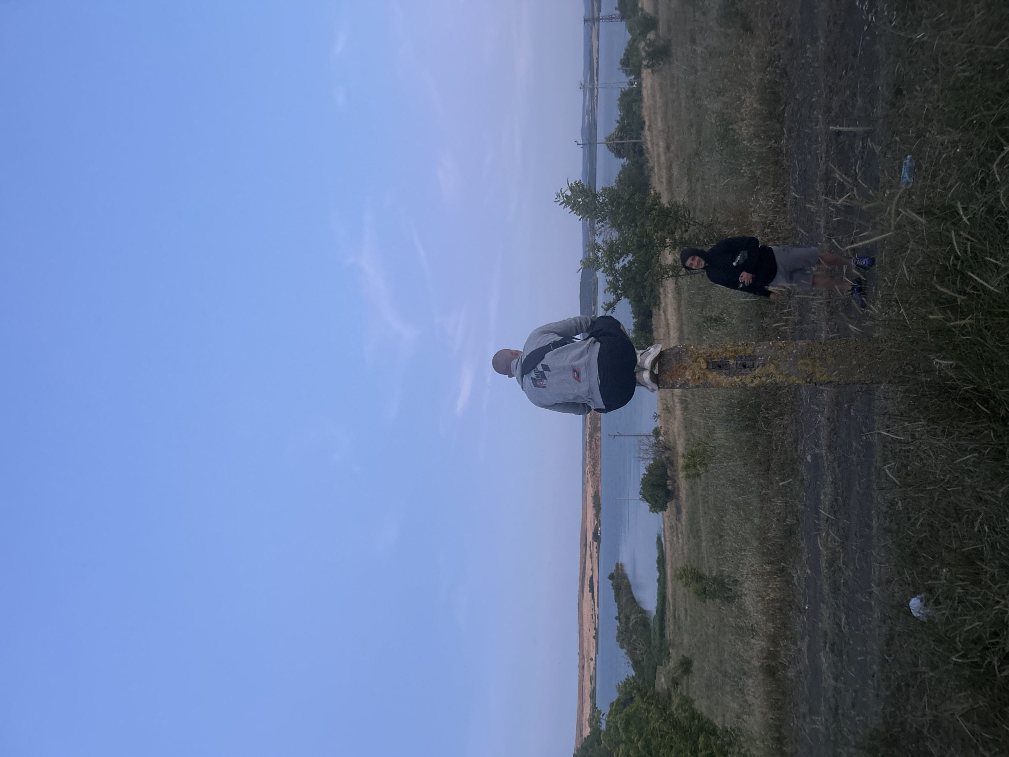 In Burgas, Bulgaria, on a dry grassy terrain with a dirt path, two individuals are present. The first individual, seated in a squatting position atop a concrete pillar, faces away from the camera towards a body of water. They are wearing a gray hooded sweatshirt and dark shorts. The second individual stands to the right of the pillar, facing the first. This person is wearing a black hooded sweatshirt and gray shorts. They are holding two clear plastic bottles. The environment includes dry grass, sparse trees, and in the background, a body of water with a distant shoreline and some infrastructure. The sky is a pale blue with faint clouds, suggesting either early morning or late evening.