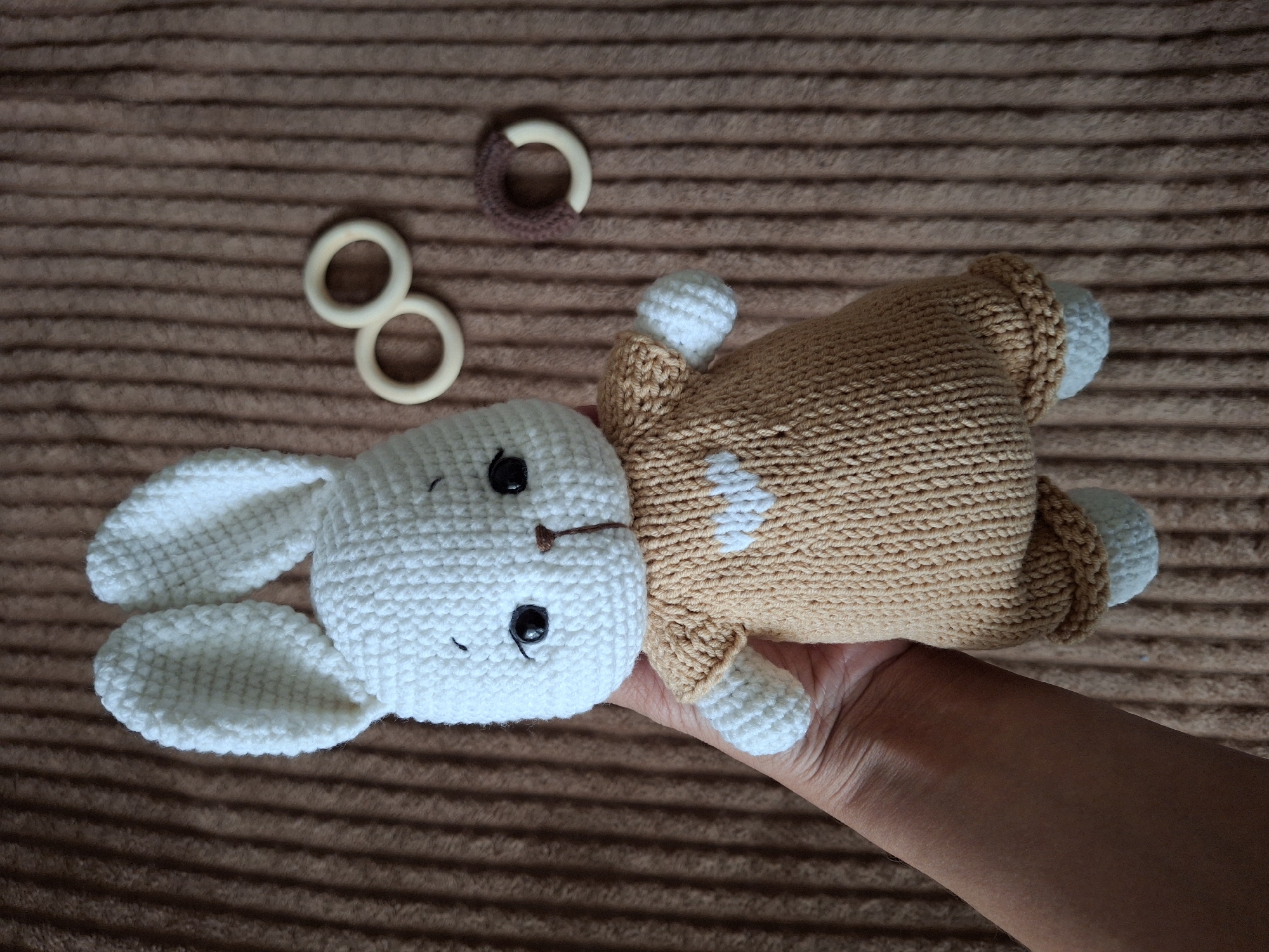 A close-up, overhead shot shows a human hand holding a crocheted bunny toy against a brown, ribbed background. The bunny has a white head with long white ears, black safety eyes, an embroidered brown nose, and a small embroidered black mouth. Its body is tan-colored with small white patches, and it has white feet and hands that emerge from tan cuffs. The hand holding the bunny is light-skinned, with visible knuckles and wrinkles, and is positioned towards the bottom right of the frame.
To the upper left of the bunny, three small rings are visible on the brown surface. Two are plain, light-colored wooden rings, while the third is a wooden ring partially covered with dark brown crochet yarn. The background is a textured fabric, possibly a blanket or rug, with distinct horizontal ridges. The lighting is soft and even, suggesting an indoor setting during daylight hours. There is no visible text in the image. The scene is likely taking place in Chaikovsky, Russia.