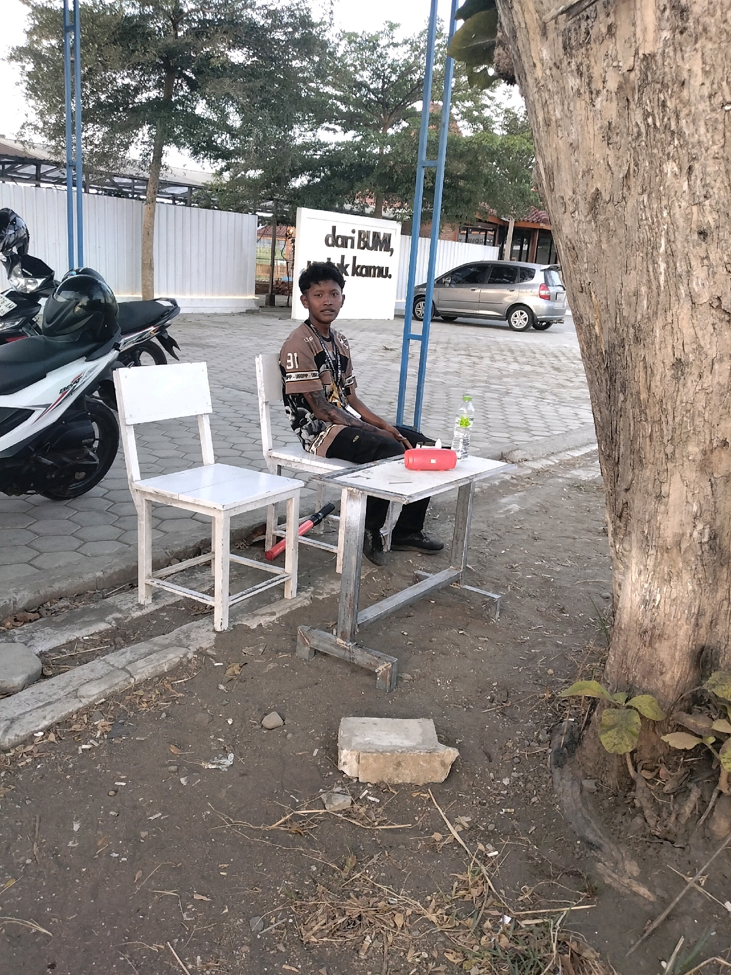 In Megulung Kidul, Indonesia, a young boy sits at a small metal table next to a white wooden chair. He is wearing a brown t-shirt with a graphic print and dark pants. On the table, a red rectangular object and a water bottle are visible. To the left of the boy, a white motorcycle is parked, and beyond it, a white wall with blue poles stands. In the background, a silver car is parked in front of a building. The scene is set outdoors on a paved area with dirt and debris scattered on the ground, next to a large tree.