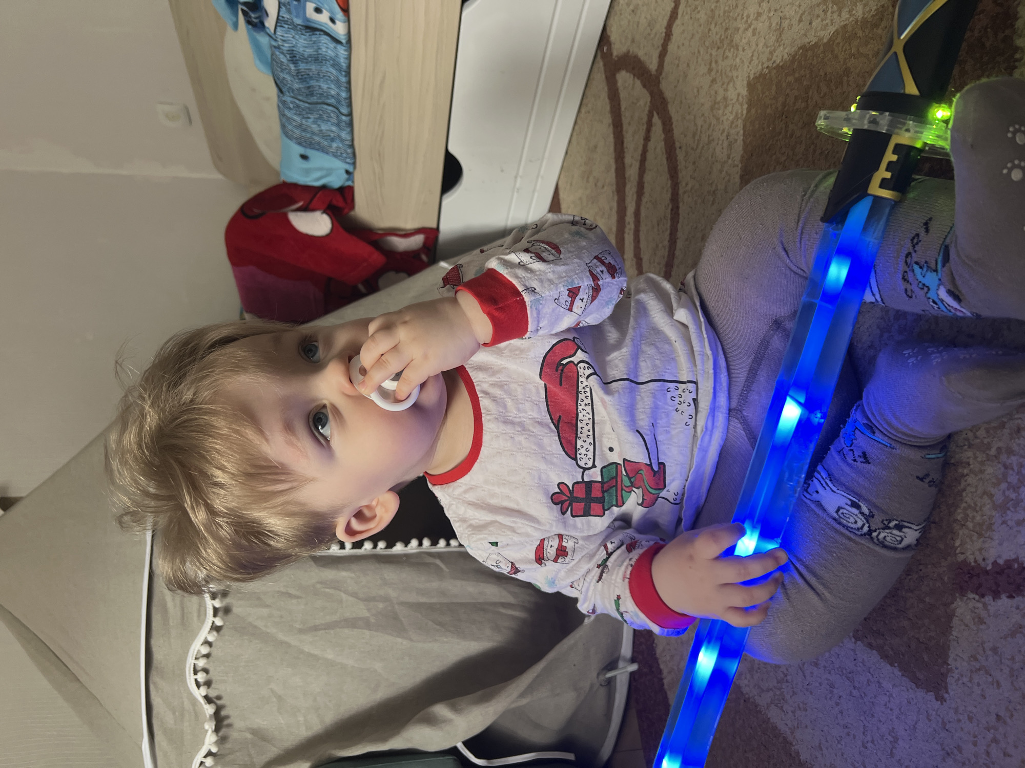 A young child, likely a toddler, is captured indoors, engaged in play in what appears to be a child's bedroom or play area in Votkinsk, Russia. The child has light blonde hair and bright blue eyes, and a white pacifier is held in their mouth. They are dressed in a festive long-sleeved pajama top, white with red cuffs and collar, adorned with Christmas motifs such as Santa Claus, gift boxes, and reindeer. The child also wears grey leggings with subtle patterns.
The child is seated or kneeling on a patterned brown rug, holding a prominent light-up toy. This toy resembles a sword or staff, emitting a vibrant blue glow along its translucent blade, while the hilt area displays distinct green lights. The child holds the blue-lit section of the toy with their left hand and appears calm and focused, looking slightly upwards and to the left of the frame.
The background shows various household items, including a segment of a light-colored wooden furniture piece, a grey fabric structure (possibly part of a playpen or small tent), and a red plush toy (possibly Spider-Man) partially visible. The indoor setting, combined with the illuminated toy, suggests a relaxed play session, potentially in the evening or a dimly lit room. No explicit text is visible in the image.
