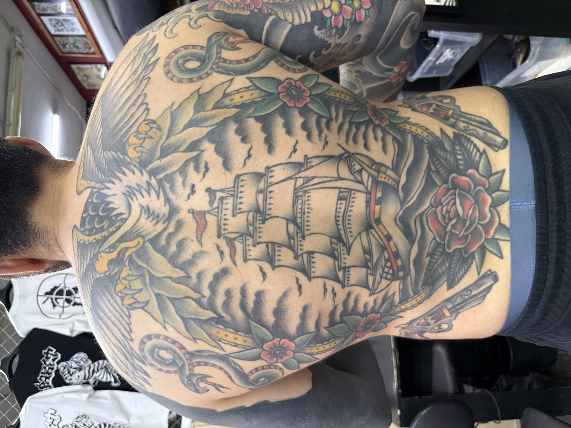 A man is seen from the back with extensive tattoos covering his upper body. The central design on his back depicts a sailing ship surrounded by clouds, a laurel wreath, and snakes. An eagle is tattooed on his left shoulder and a gun and rose on his right side. The man is wearing black athletic shorts with a blue waistband. The environment appears to be indoors, possibly a tattoo studio, with t-shirts visible in the foreground and assorted items in the background. No evidence suggests Moscow, Russia.