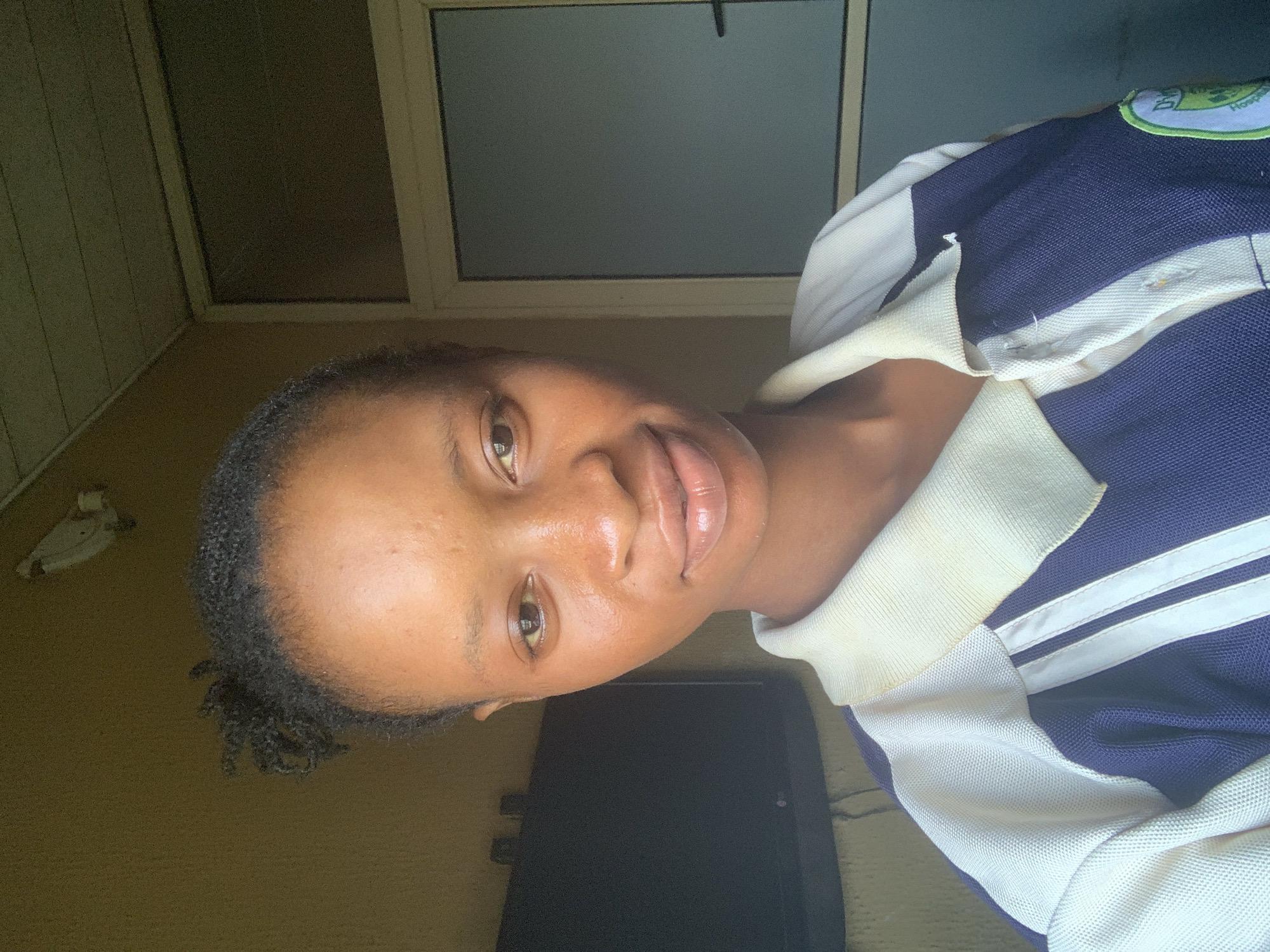A young Black woman with dark hair pulled back is shown in a close-up, self-taken photograph. She is wearing a collared shirt with navy blue and off-white panels, and white stripes on the sleeves. Her skin is a rich brown, and she has a subtle smile. In the background, a dark television screen is visible to the left, and a closed glass door with a handle is on the right. The wall behind her is a textured, yellowish-brown color, and the ceiling is visible at the top of the frame. The lighting suggests the photo was taken indoors, possibly in a residential setting, given the appearance of the wall and door. The location is identified as Shagamu, Nigeria.