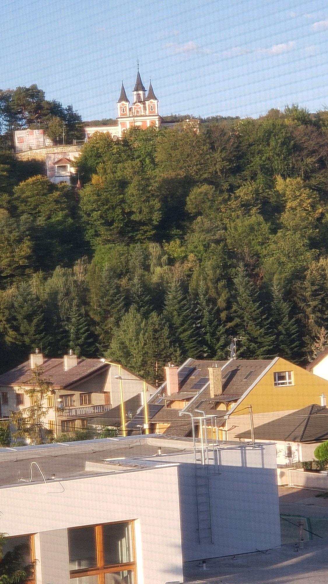 The image depicts a view of the Church of the Holy Trinity in Prešov, Slovakia, as seen from a distance. The church, characterized by its red and white facade and multiple spires, is perched atop a verdant, tree-covered hill. In the foreground, several residential buildings with pitched roofs are visible. These structures exhibit a range of architectural styles and colors, with one building painted yellow and another in shades of beige and brown. A white, modern-looking building with large windows occupies the lower portion of the frame. A metal ladder is affixed to the side of this building, extending upwards. The sky above is a clear blue with a few wispy clouds. The overall scene suggests a residential area in Prešov, with the church serving as a prominent landmark on the skyline.