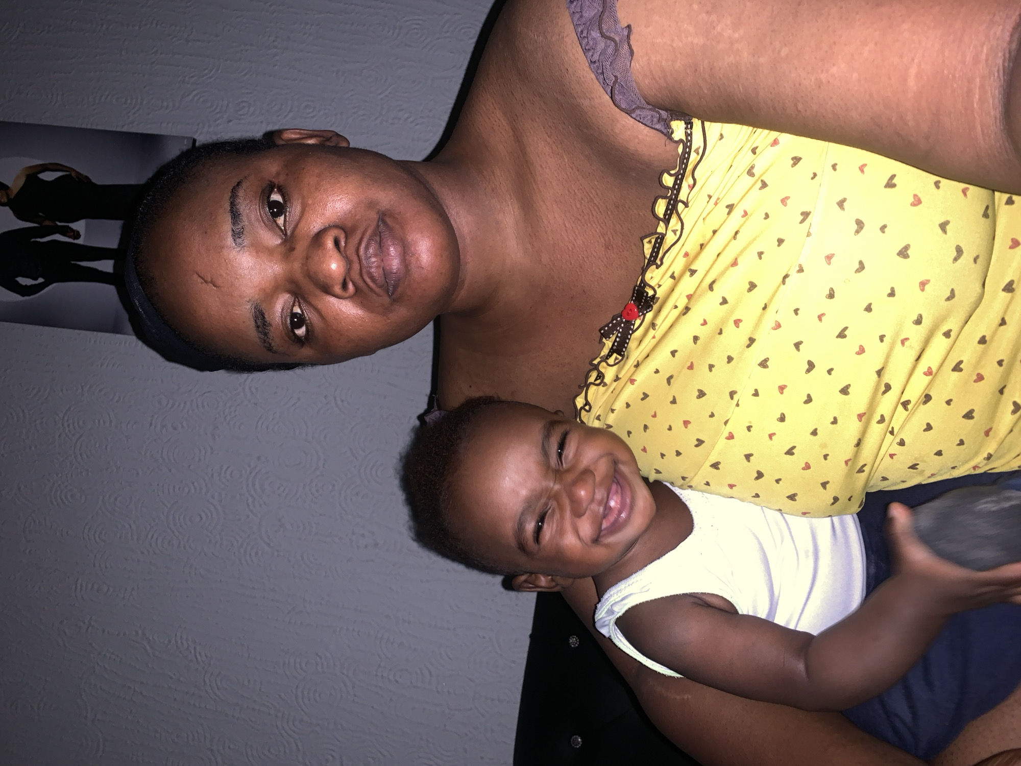 A medium shot shows a woman and a child indoors. The woman, with dark skin and short hair, wears a yellow top with a heart pattern. Her expression is neutral, and her gaze is directed towards the camera. The child, also with dark skin, sits on the woman's lap, wearing a white tank top and blue shorts. The child is smiling widely, with eyes squinted, and is looking towards the camera. A framed portrait of a couple is visible on the wall behind them. The environment appears to be a residential interior, possibly in the city of Barakin Akawo, Nigeria.