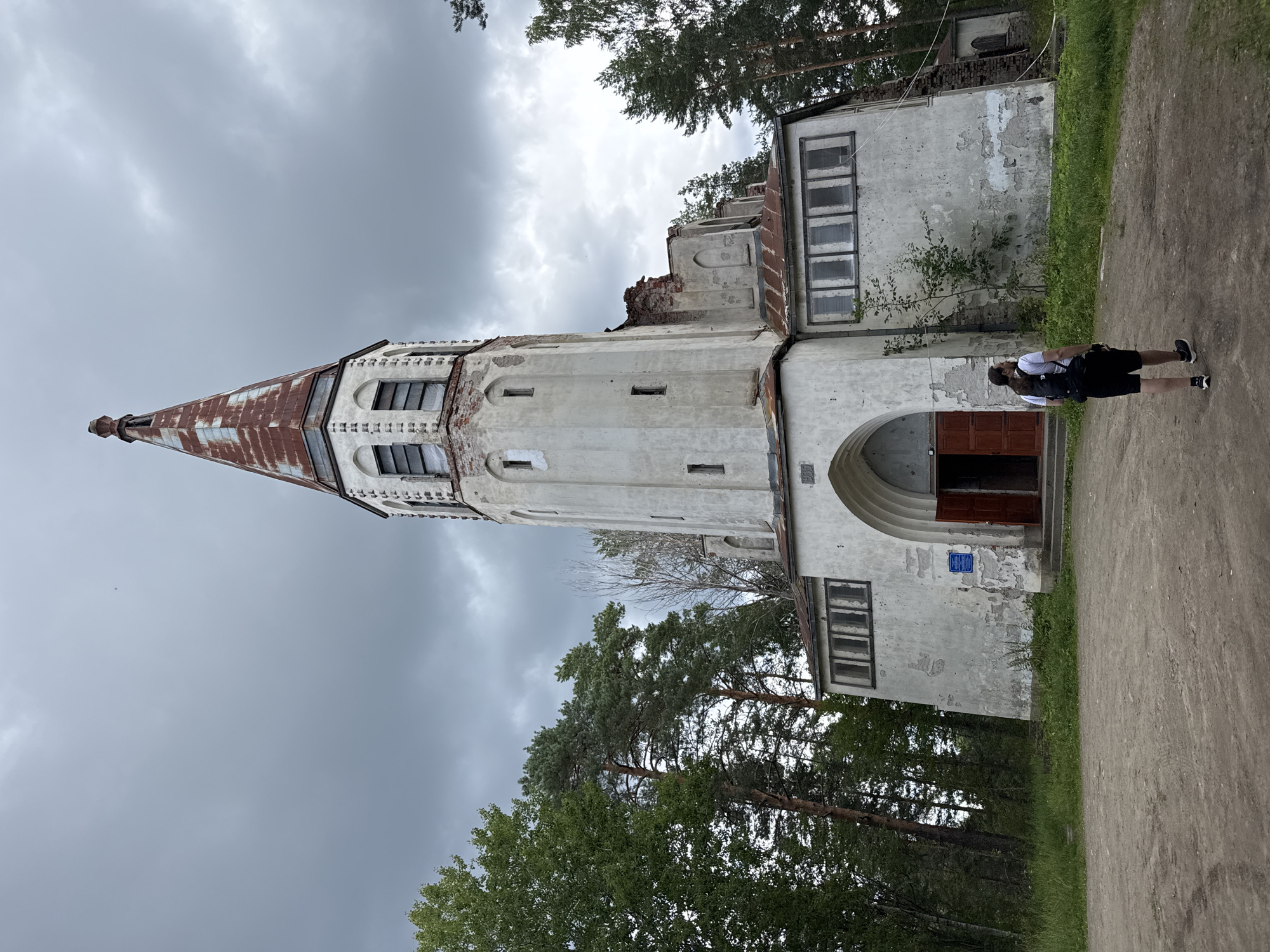 In Lumivaara, Russia, a person with a backpack walks away from a dilapidated white church with a rusty, conical roof. The church features arched doorways and windows, some of which are broken or boarded up. The building is partially in ruins, with exposed brickwork and peeling paint. Surrounding the church are tall trees, including pines, and a cloudy, overcast sky looms above. The ground in front of the church is a dirt path.