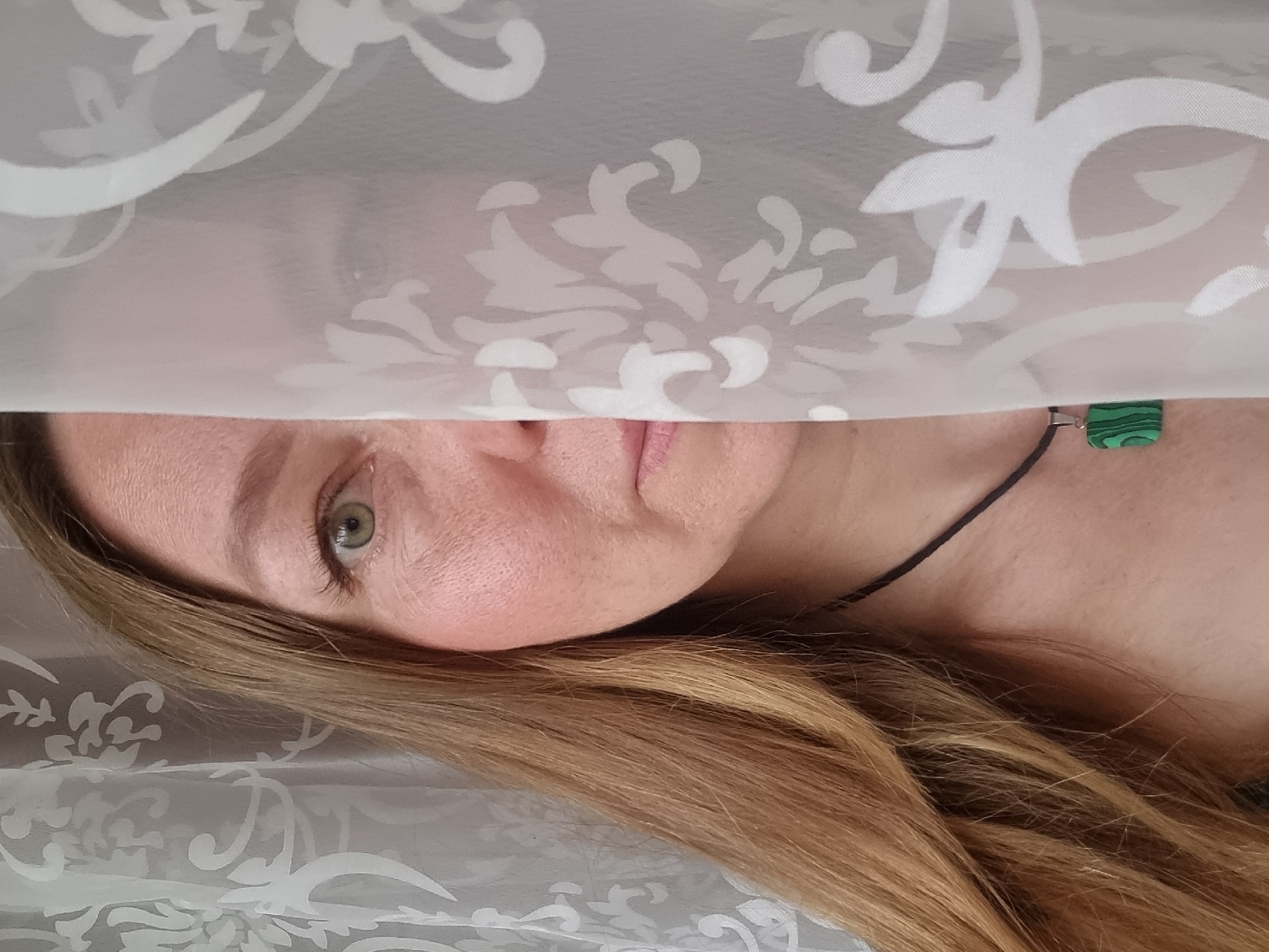A person's face is partially obscured by a patterned, semi-transparent fabric. The person has fair skin, blonde hair, and a green eye is visible. A black cord necklace with a green and black geometric pendant is visible on their neck. The fabric overlaying the face features a repeating floral or damask pattern in white and gray tones. No information about the city of Chaikovsky, Russia is discernible from the image.