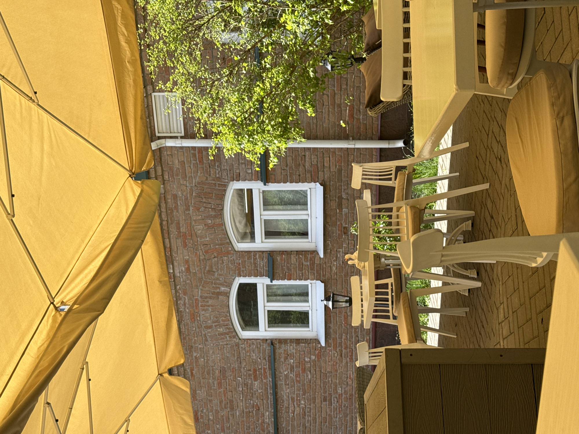 The image depicts an outdoor dining area, likely a café or restaurant, in Penza, Russia. The focus is on a brick building with two arched windows and a smaller, rectangular window above. A yellow umbrella shades the seating area, which consists of white tables and chairs arranged on a paved surface. A streetlight is visible between the windows. Green foliage from trees obscures parts of the building and provides additional shade. The scene suggests a warm day in a European city.
