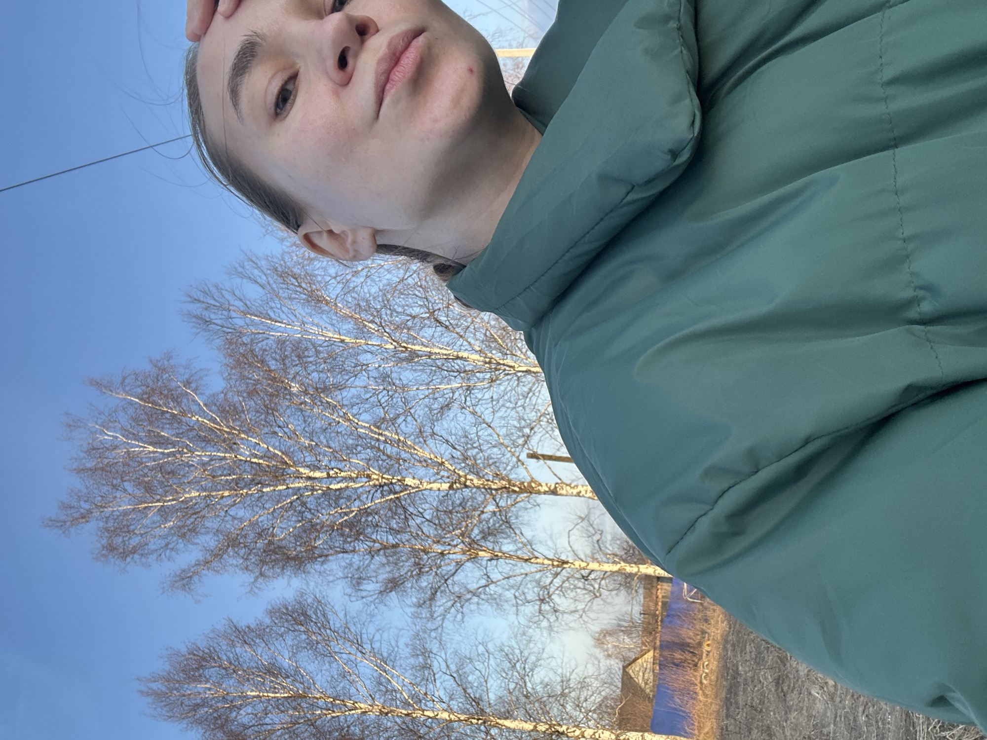 The image depicts a woman, possibly in her late teens or early twenties, wearing a green puffer jacket, standing outdoors. The background shows leafless trees against a blue sky. In the distance, there is a glimpse of a building, likely residential, with construction materials in its yard. A powerline is visible against the sky. The woman is holding her hand to her forehead. Without specific locational markers, it's impossible to definitively confirm the location as Kharnavy, Russia.