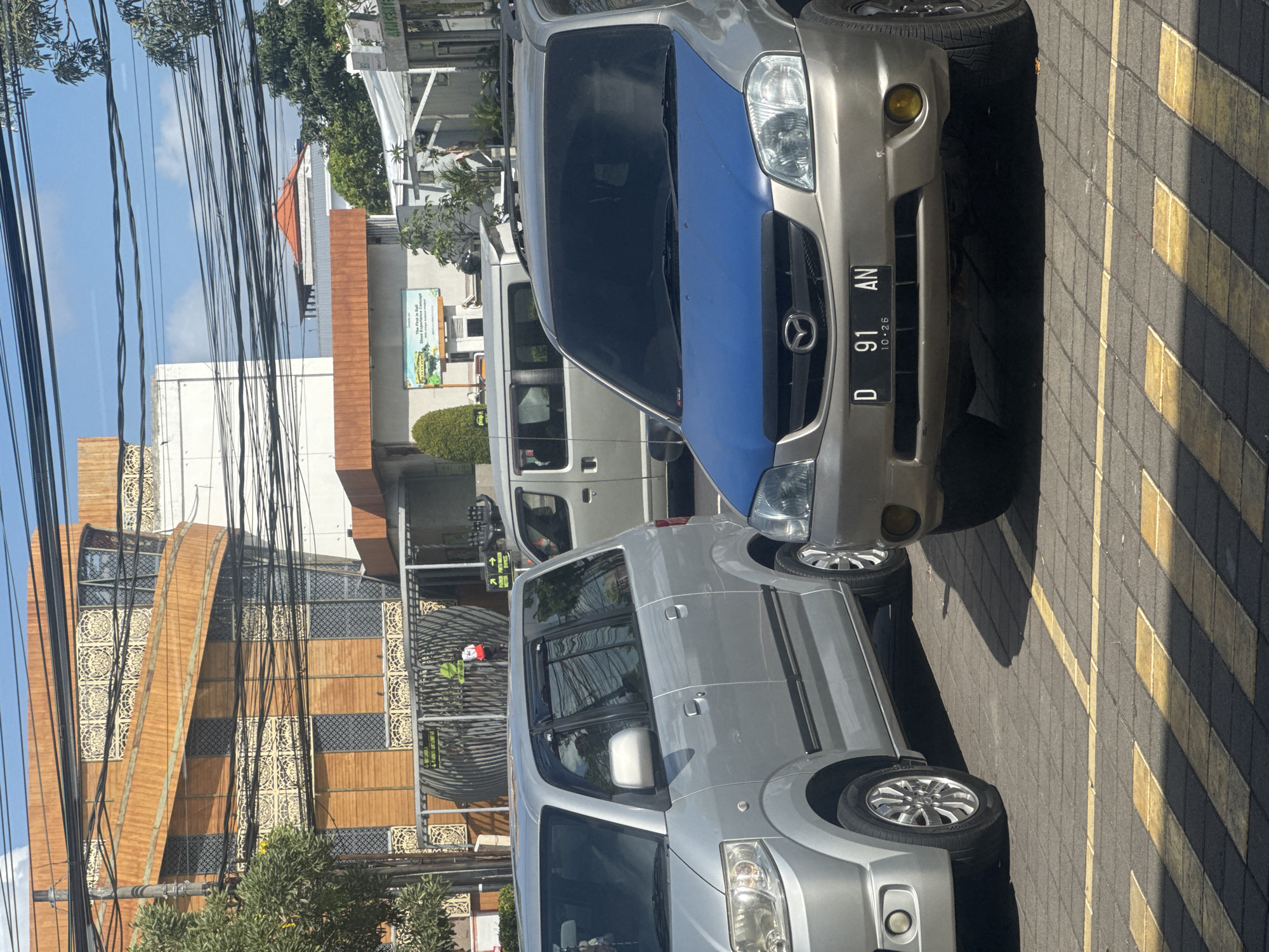 The image is a medium shot of two cars parked on a paved lot in Kuta, Indonesia. The car in the foreground is a tan SUV with a blue hood, and its license plate reads "D 91 AN 10.26". To its left is a silver minivan. In the background, a modern building with wooden accents and intricate metalwork can be seen, along with overhead electrical wires and trees. The ground is paved with interlocking bricks, and yellow lines mark parking spaces. A person wearing a green shirt and a backpack is visible in the background near a gate.