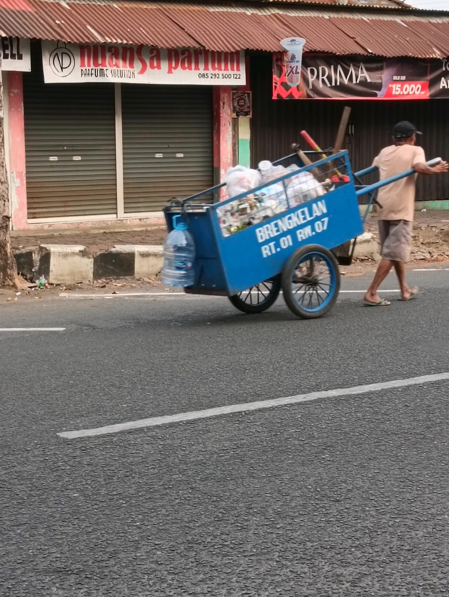 A man in a tan shirt and shorts pushes a blue, metal-framed cart laden with trash on a paved road in Purworejo, Indonesia. The cart has two wheels and a long handle. A large blue plastic water bottle is attached to the side of the cart. The man is walking away from the camera and appears to be in motion. Behind him, a building with a corrugated metal roof has signs that read "NUANSA PARFUM" and "XL PRIMA" with a price of "15.000". The building has metal roll-up doors. The road has a white line painted on it. The scene appears to be during daylight hours.