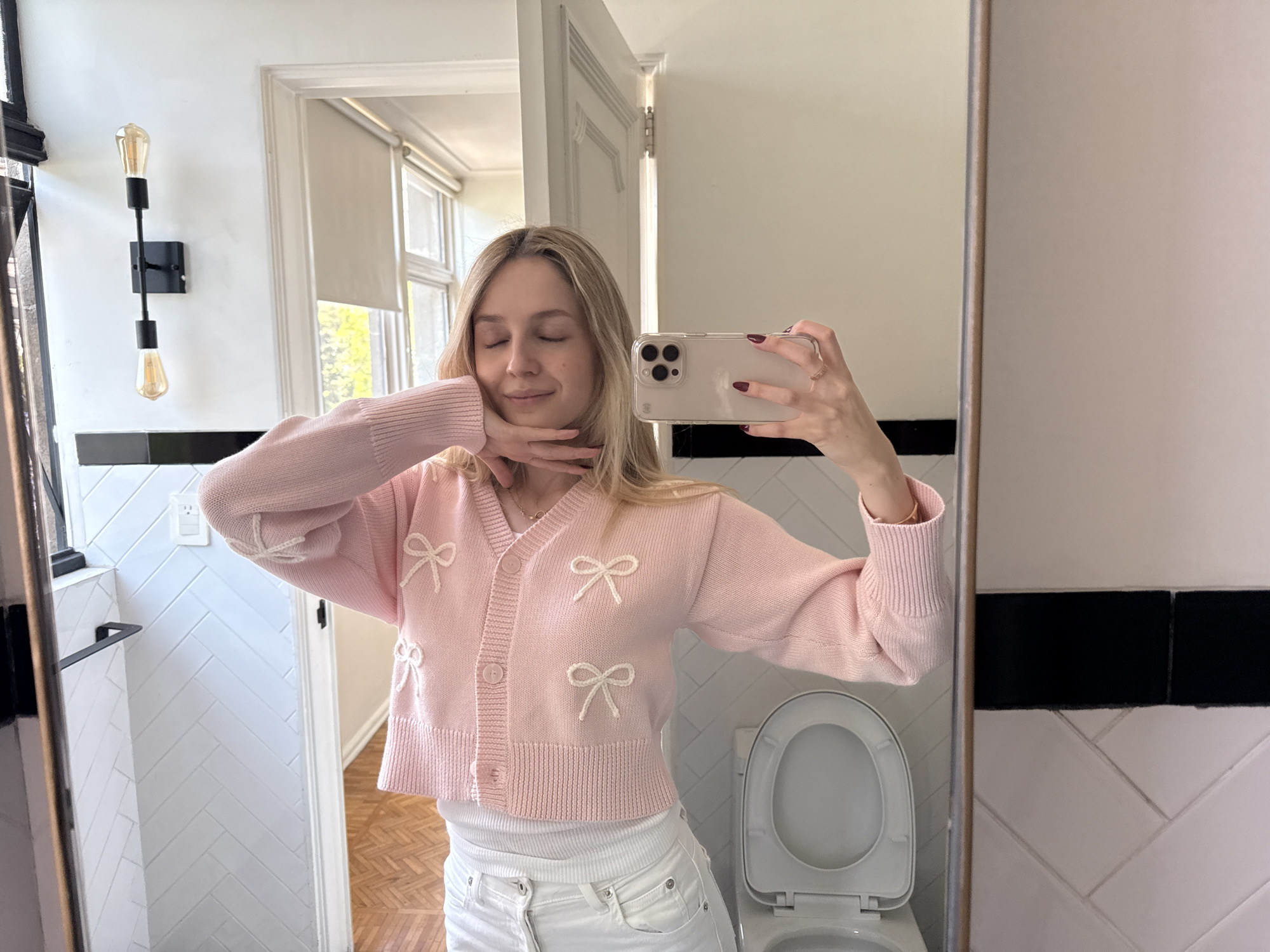A young adult female is depicted taking a mirror selfie in a bathroom located in Mexico City, Mexico. She has blonde hair and is shown with her eyes closed, displaying a subtle smile. Her left hand is positioned gently beneath her chin. She is wearing a pink cropped cardigan adorned with white bow appliqués, a white tank top, and white jeans. Her fingernails are painted with dark red polish. She holds a light-colored smartphone, fitted with a clear protective case, in her right hand, which is visible as a reflection.
The background, primarily observed through the mirror's reflection, consists of white rectangular wall tiles arranged in a herringbone pattern on the lower half, topped by a horizontal black border tile. Above this, the walls are painted white. A black wall-mounted sconce with two exposed Edison-style filament bulbs is installed on the left wall. A white toilet with its lid open is situated in the lower right portion of the scene. A black horizontal towel bar is visible on the left wall. In the upper left background, a white doorway with a closed door featuring vertical paneling is partially visible. Beyond the doorway, a window with a partially drawn white roller blind reveals green foliage outside. The flooring appears to be light-colored wood with a parquet or patterned design.