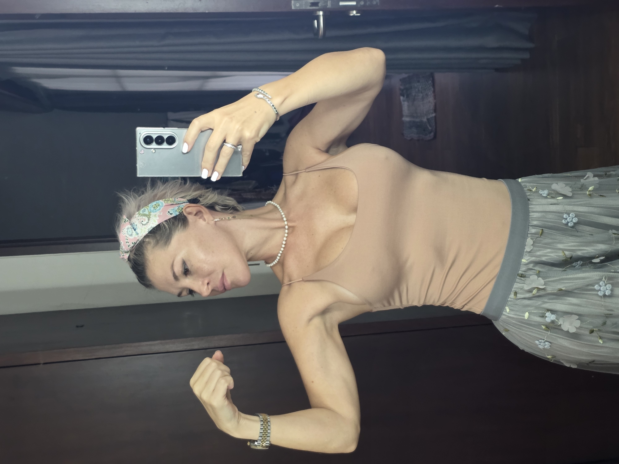 In North Kuta, Indonesia, a woman is posing in what appears to be a bedroom or dressing area, reflected in a mirror. She is wearing a beige spaghetti strap top and a grey skirt with floral embroidery. She has blonde hair styled in a messy bun, a pearl necklace, earrings, a watch on her left wrist, and a bracelet and ring on her right hand. The woman is flexing her left bicep and holding a smartphone in her right hand, likely taking the selfie. The background shows a dark wooden door, a curtain, and a rug on the floor.