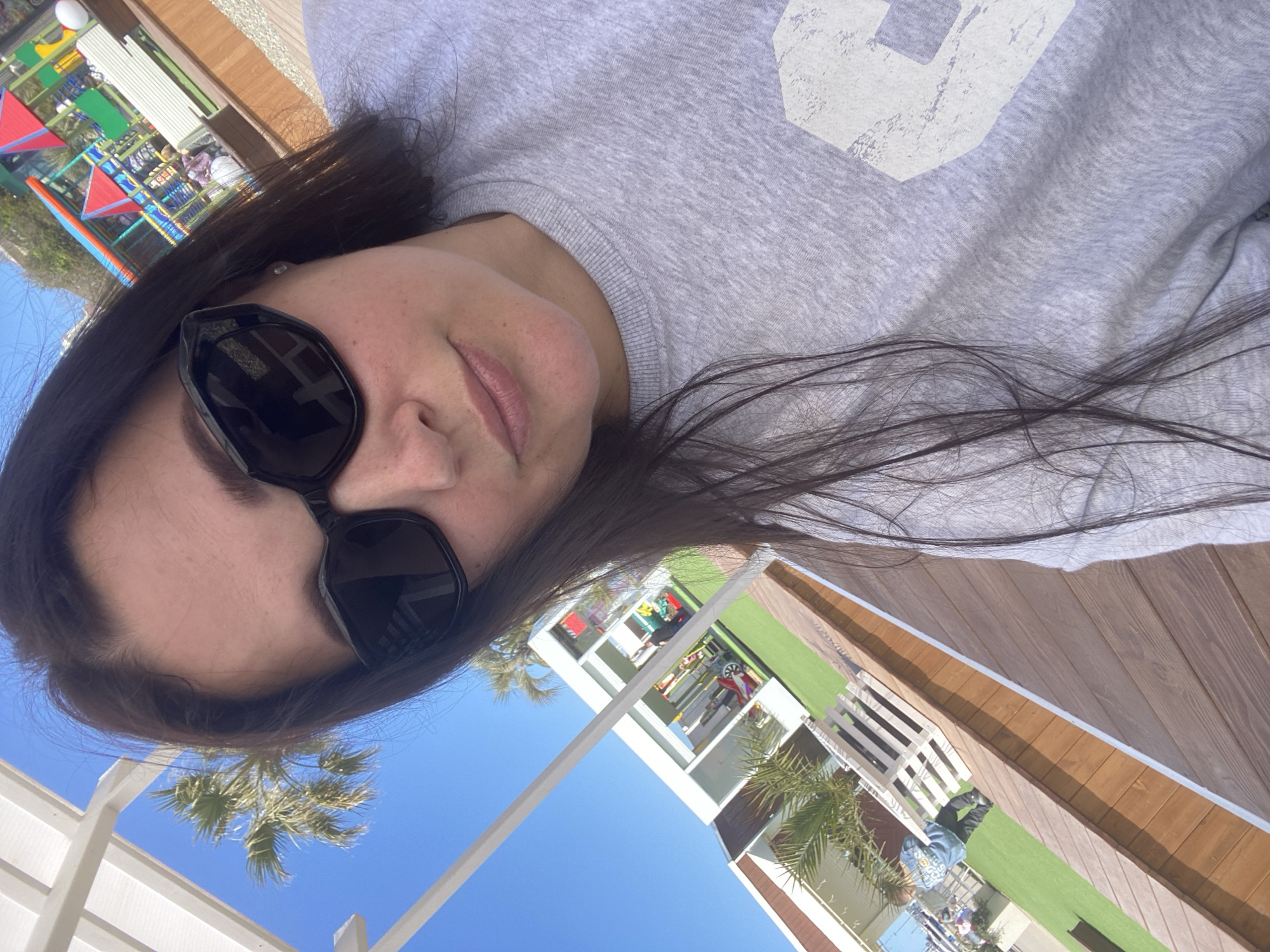 A selfie-style, slightly angled close-up shows a woman with dark hair wearing sunglasses and a grey sweatshirt. The background reveals an outdoor scene with a clear blue sky visible behind a white architectural structure. Palm trees are also visible. In the blurred background, there appears to be a colorful playground and a multi-story building. Given these features, the photo was likely taken in a resort area with a playground and palm trees. While it's impossible to definitively confirm without additional information, the description matches common features of resorts in coastal cities like Sochi, Russia.