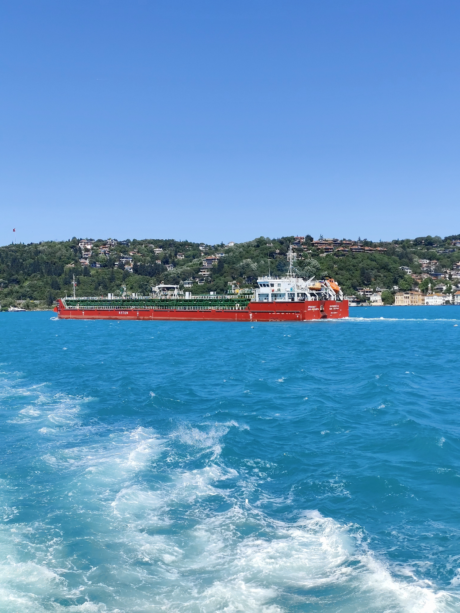 A full-length image of a red cargo ship on the water, likely the Bosphorus, in the Sarıyer district of Istanbul, Turkey. The ship, visible in its entirety, appears to be a tanker, with green pipelines running along its deck. The body of water surrounding the ship is a clear, bright turquoise, with white frothy ripples visible in the foreground of the image, presumably from the wake of another boat. The sea meets a landmass in the background, covered in dense green vegetation and residential buildings. The sky above is clear and cloudless. A small red flag is visible in the distant, upper left corner.