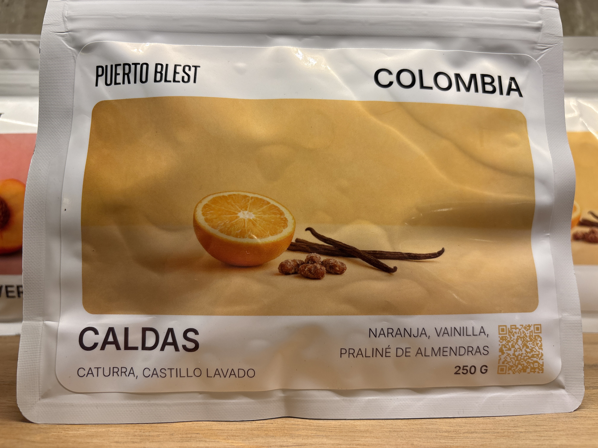 The image shows a white, sealed bag of Puerto Blest coffee from Colombia. The Caldas region is specified, along with the coffee varieties Caturra and Castillo Lavado. The flavor profile listed is orange, vanilla, and almond praline. The bag contains 250 grams of coffee. A photograph of an orange slice, vanilla bean, and sugared almonds are featured. A QR code is printed on the lower right side of the bag. The bag is placed on a wooden surface.