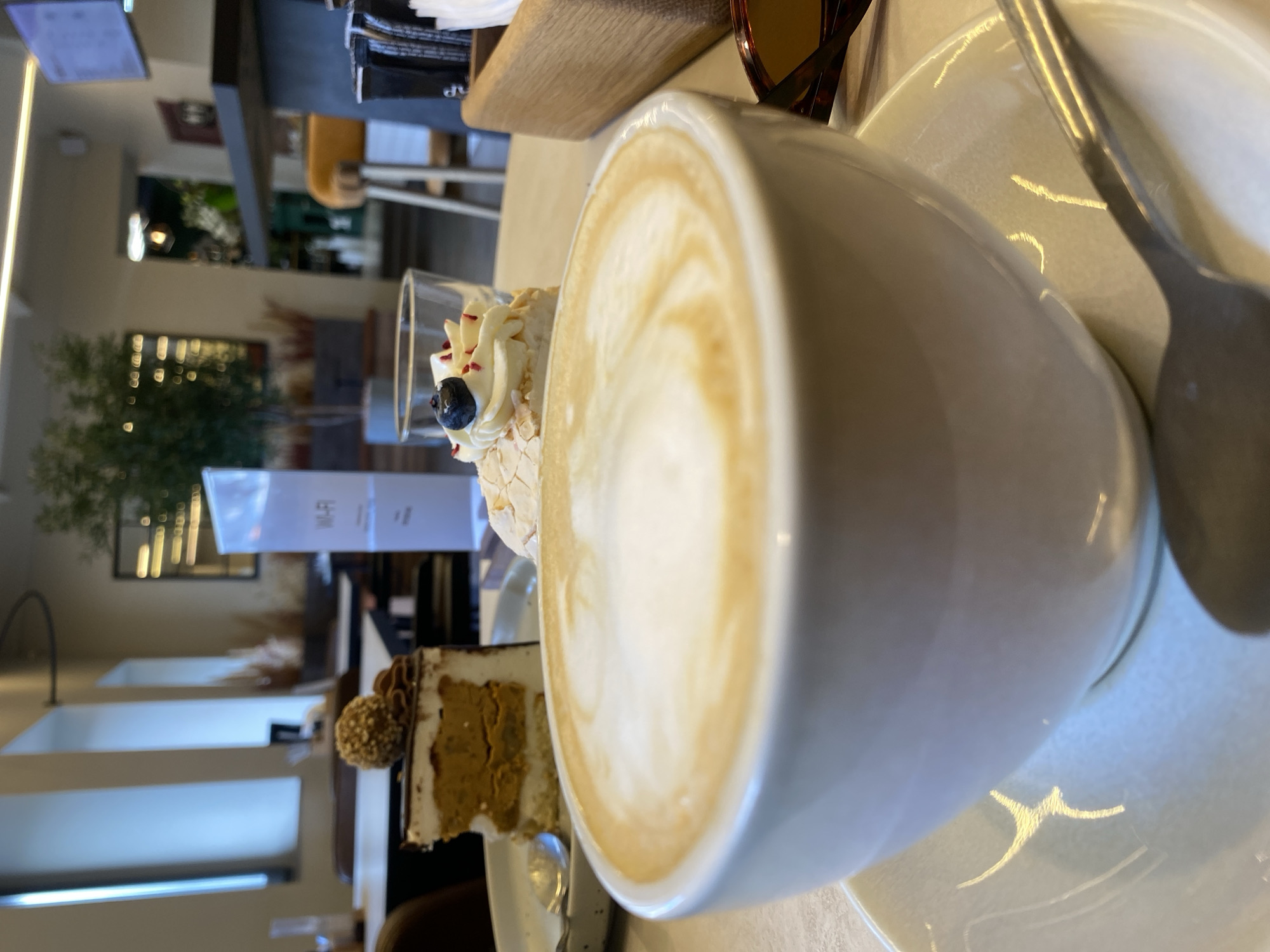 A close-up shot captures a white ceramic cup of cappuccino, filled to the brim with frothy milk art, placed on a matching saucer with a spoon resting beside it. In the background, a slice of layered cake with chocolate ganache and a hazelnut topping is visible on a plate, alongside a delicate dessert topped with whipped cream, a blueberry, and sprinkles. A glass of water is also present. A white sign with "Wi-Fi" printed on it stands upright. The setting appears to be a bright, modern cafe, with tables, chairs, and plants visible. The overall atmosphere suggests a relaxed moment with dessert and coffee. This image is not associated with Votkinsk, Russia.