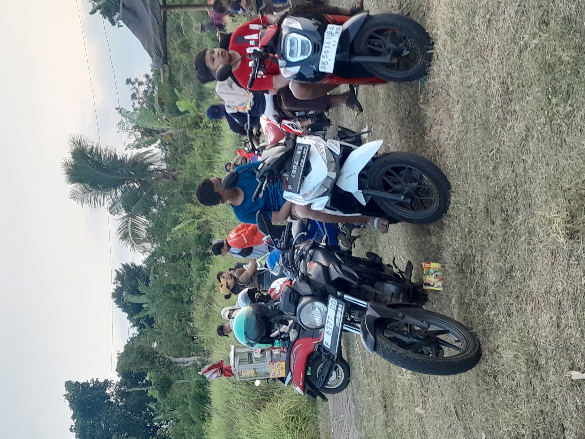 The image depicts an outdoor scene in Genteng, Indonesia, likely a public gathering or market area, characterized by the presence of several motorcycles parked on dry, grassy ground. In the foreground, a black motorcycle with license plate "P 6177 DQ" is prominently displayed. To its right, a white scooter with license plate "P 4982 XS" is visible, with a young boy in a blue t-shirt sitting on it. Further to the right, a red and black scooter with license plate "DK 5614 UBA" is parked, with another young boy in a red t-shirt seated on it.
In the background, a red scooter is parked near a small food stall with a visible menu. A variety of people are scattered throughout the scene, some standing and conversing, others seemingly observing the surroundings. The environment is lush with green vegetation and trees, suggesting a rural or semi-rural setting within Genteng. The sky is overcast, indicating a cloudy day. The overall impression is of a casual, community-oriented gathering, possibly a local market or event.
