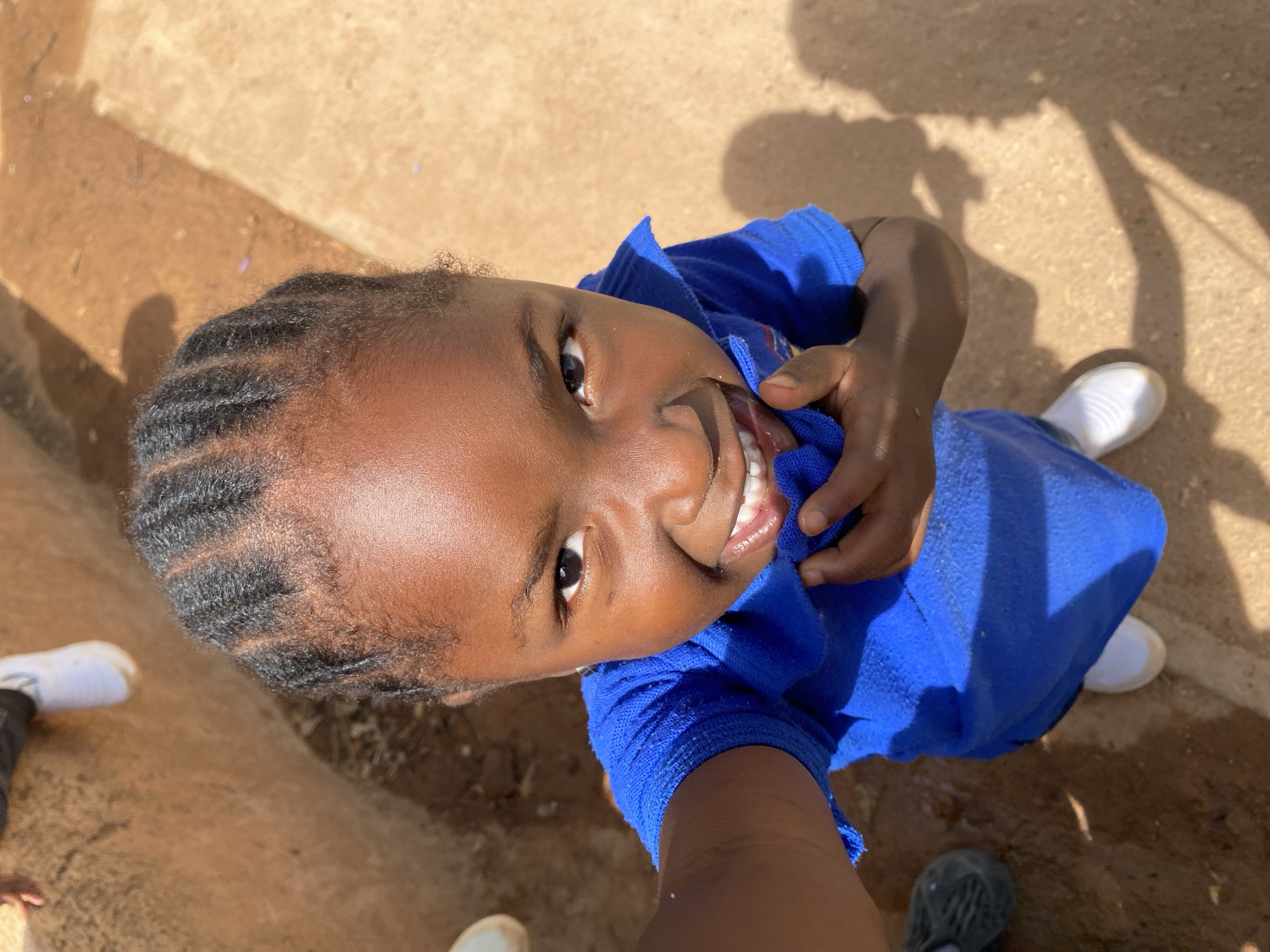 A close-up, upward-facing shot shows a young Black girl with braided hair, smiling and holding a blue garment to her chest. She is wearing white sneakers. The location is Gura Top, Nigeria, indicated by the earthy, reddish-brown ground underfoot. Shadowy figures, presumably of other people, are visible to the upper right. One person is wearing black sneakers. The photograph is taken outdoors in daylight.