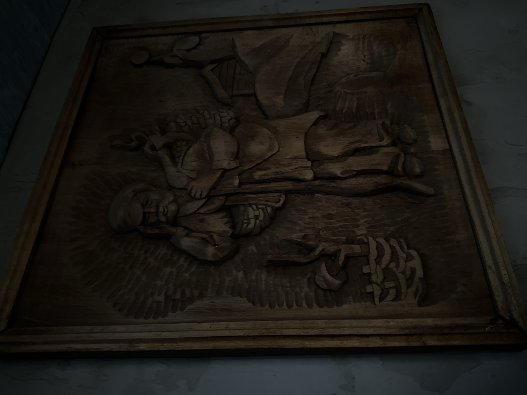 The image shows a carved wooden relief depicting a man dressed in traditional clothing, possibly a farmer or traveler, carrying a basket over his shoulder and holding a walking stick. He is shown in a rustic setting, with a small house and a basket of produce at his feet. The carving is framed by a wooden border. The artwork is mounted on a wall that appears to be textured and painted a light color. The lighting is dim, casting shadows and highlighting the intricate details of the carving. The location is specified as Luhansk, Ukraine.