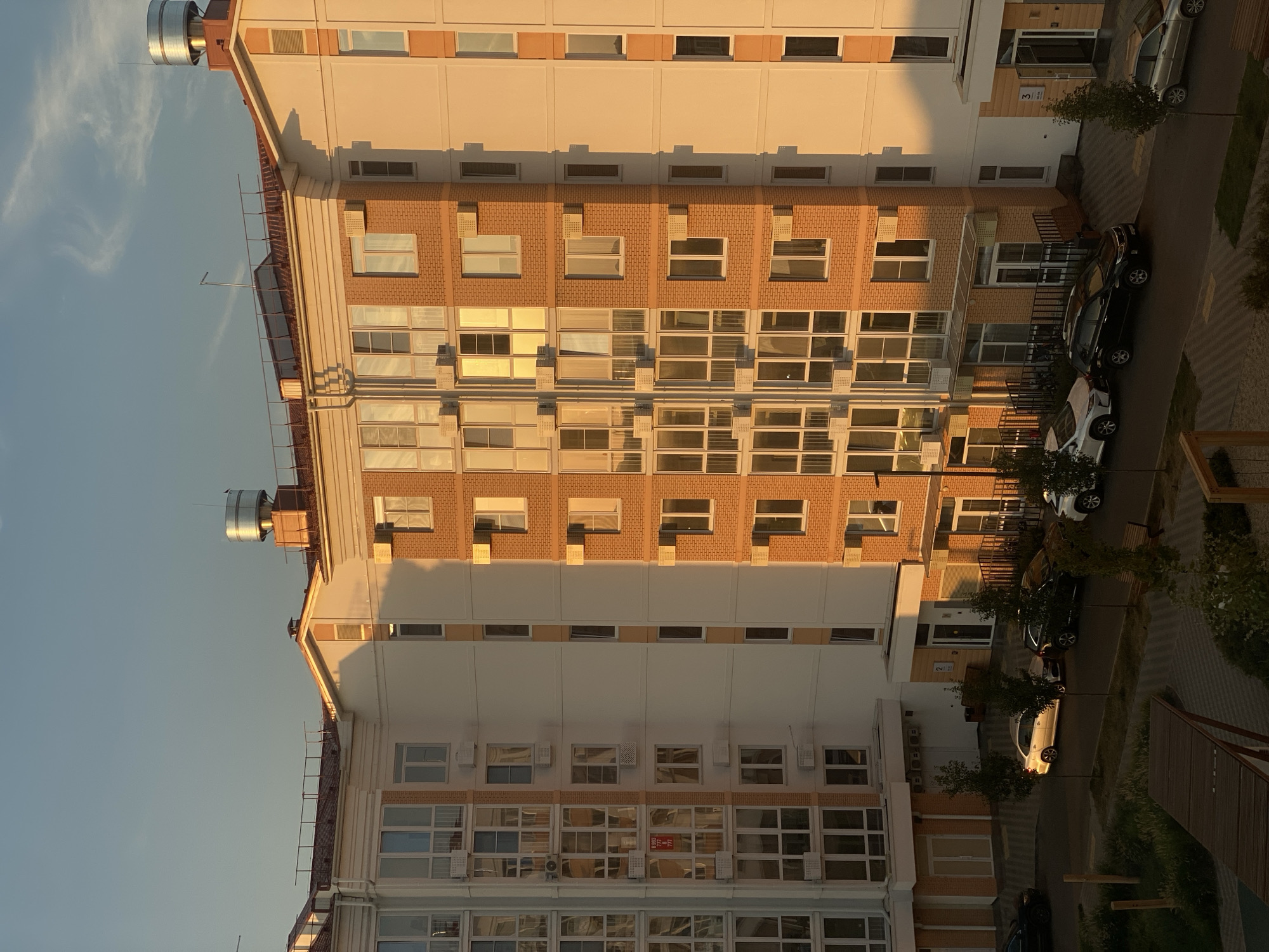 This is a photograph of a residential building in Stopyatyy, Russia, taken during sunset. The building is a modern, multi-story structure with a facade of light-colored stucco and orange brick. The windows are large and numerous, reflecting the warm glow of the setting sun. Several cars are parked in the courtyard below, and a few trees dot the landscape. The sky is clear, with a few wispy clouds visible. The scene is peaceful and serene, bathed in the golden light of dusk.