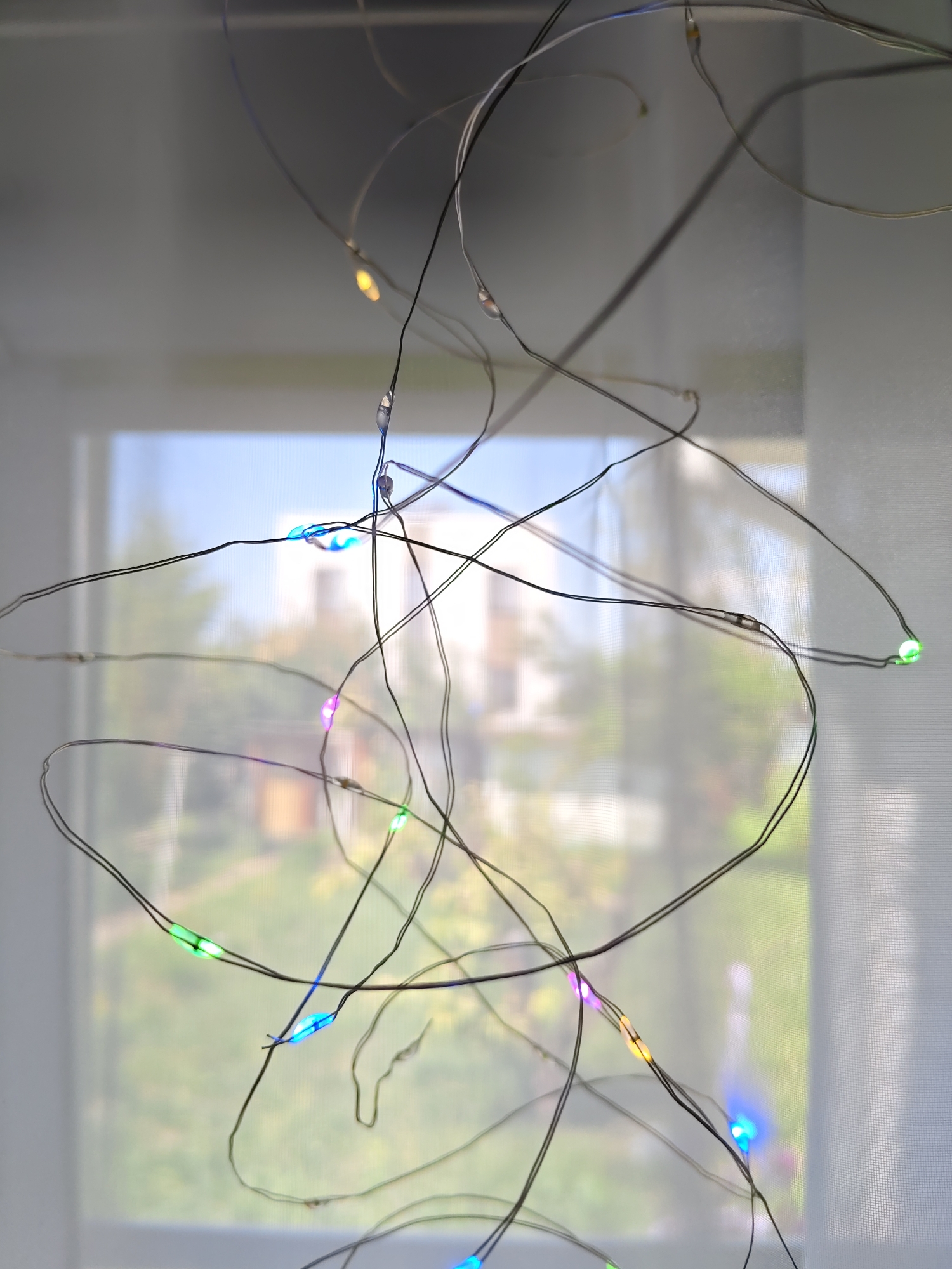 A close-up, vertical, indoor shot captures a tangle of thin, dark wires with small, multi-colored LED lights strung along them. The lights are glowing in various colors, including blue, green, purple, and yellow. The wires are draped across the frame, obscuring a blurry background. The background appears to be a window with sheer white curtains, through which an outdoor scene is visible. The outdoor scene is out of focus but suggests trees, buildings, and a bright sky, possibly indicating daytime. The image does not provide any specific landmarks or visual cues to pinpoint its location within Rostov-on-Don, Russia.