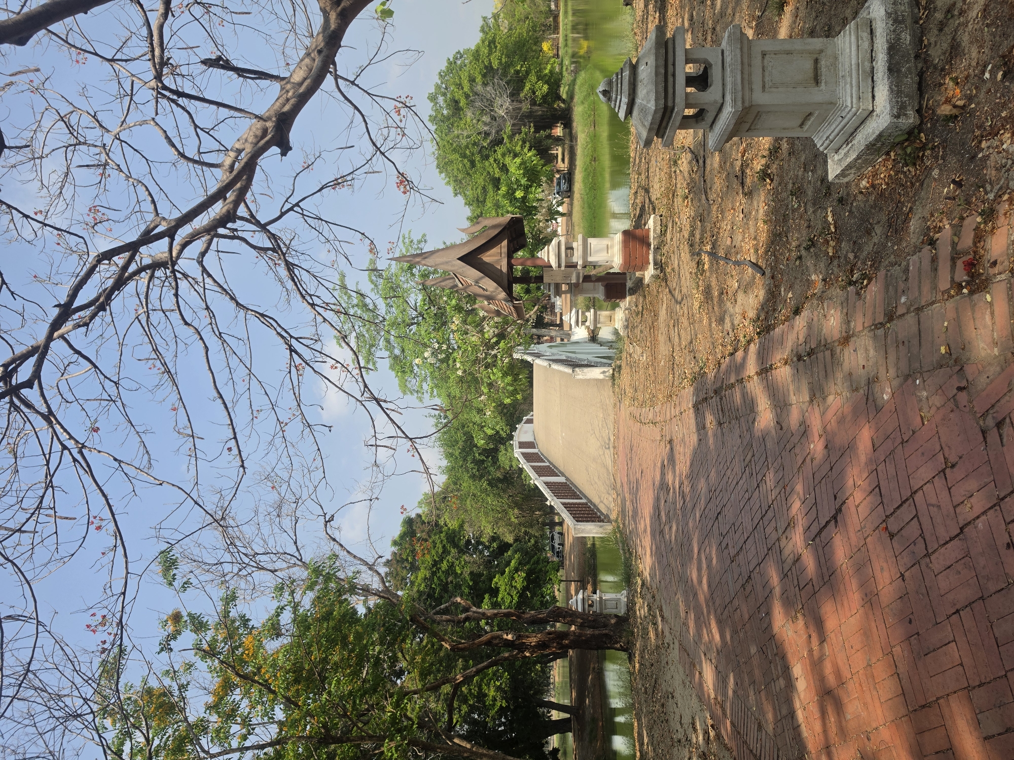 The scene depicts a park in Phra Nakhon Si Ayutthaya City Municipality, Thailand. A brick walkway leads towards a small Thai-style pavilion on a pond. Stone lanterns line the walkway. Trees are on either side, some with bare branches. The sky is partially visible through the branches.
