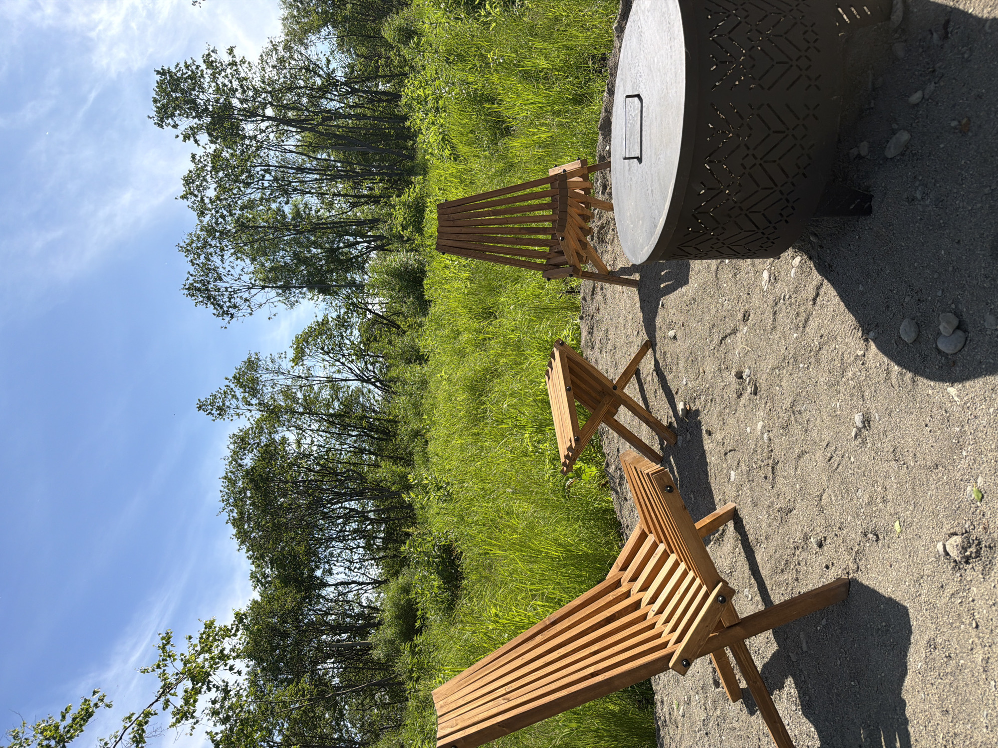 A sunlit outdoor scene features two wooden chairs and a small wooden table arranged on a sandy area. In the foreground, a large, dark circular fire pit with a decorative patterned base sits to the right. Behind the furniture, a dense area of tall, green grass and trees stretches across the background under a clear blue sky with sparse white clouds. The image appears to be taken during the daytime, likely in a natural or park-like setting.