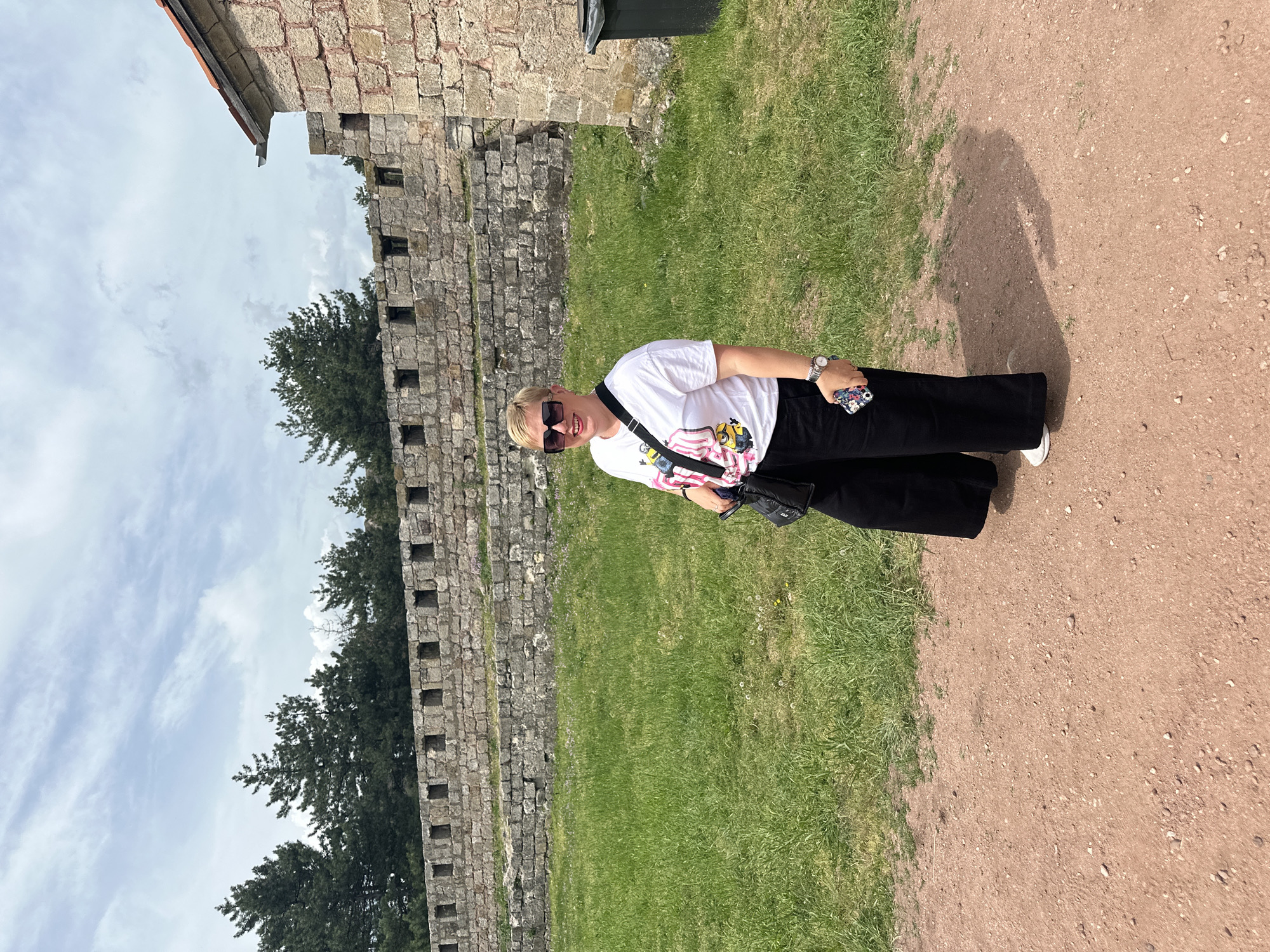 The image depicts a woman standing outdoors. She has short blonde hair and is wearing sunglasses, a white t-shirt with a graphic design, black pants, and white shoes. She has a crossbody bag and holds a phone in her right hand. The setting appears to be near the Belogradchik Fortress in Belogradchik, Bulgaria, indicated by the prominent stone wall in the background. The ground consists of grass and a dirt path. The sky is visible in the upper left corner and is light blue with some clouds.
