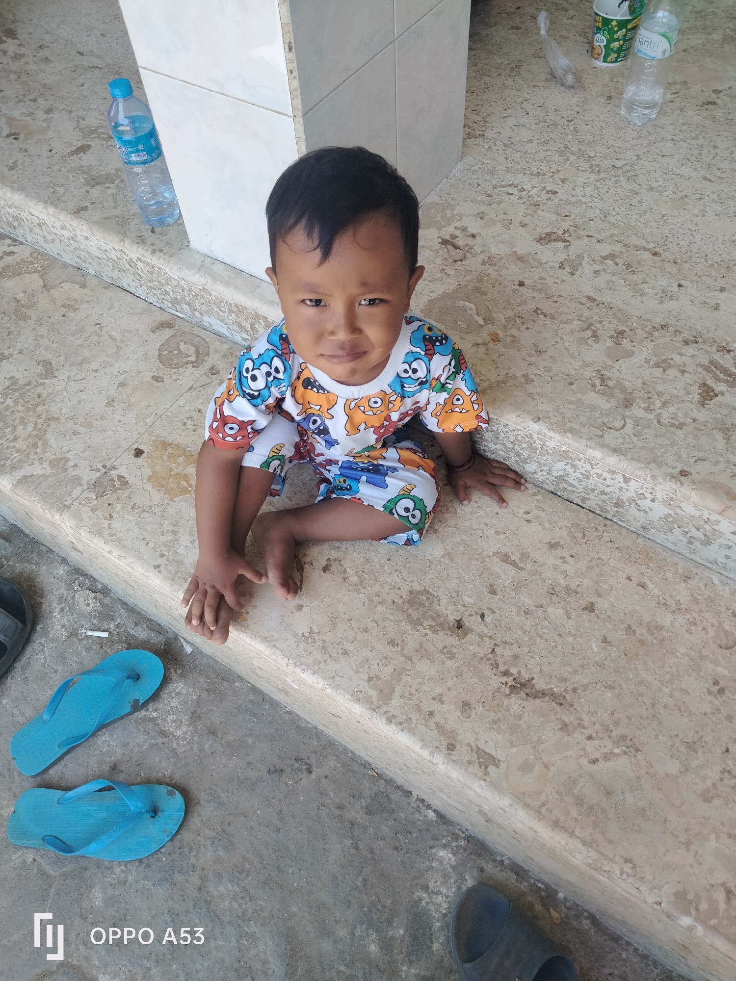 A young child with dark hair is sitting on a concrete step outside. The child is wearing a t-shirt and shorts with a cartoon monster pattern. The child is looking at the camera with a neutral expression. There are two blue flip-flops on the ground in front of the child. To the left of the child, there is a plastic bottle of water with a blue cap. In the background, there is a white tiled wall and a patterned floor. The image appears to have been taken in the city of Badung, Indonesia.