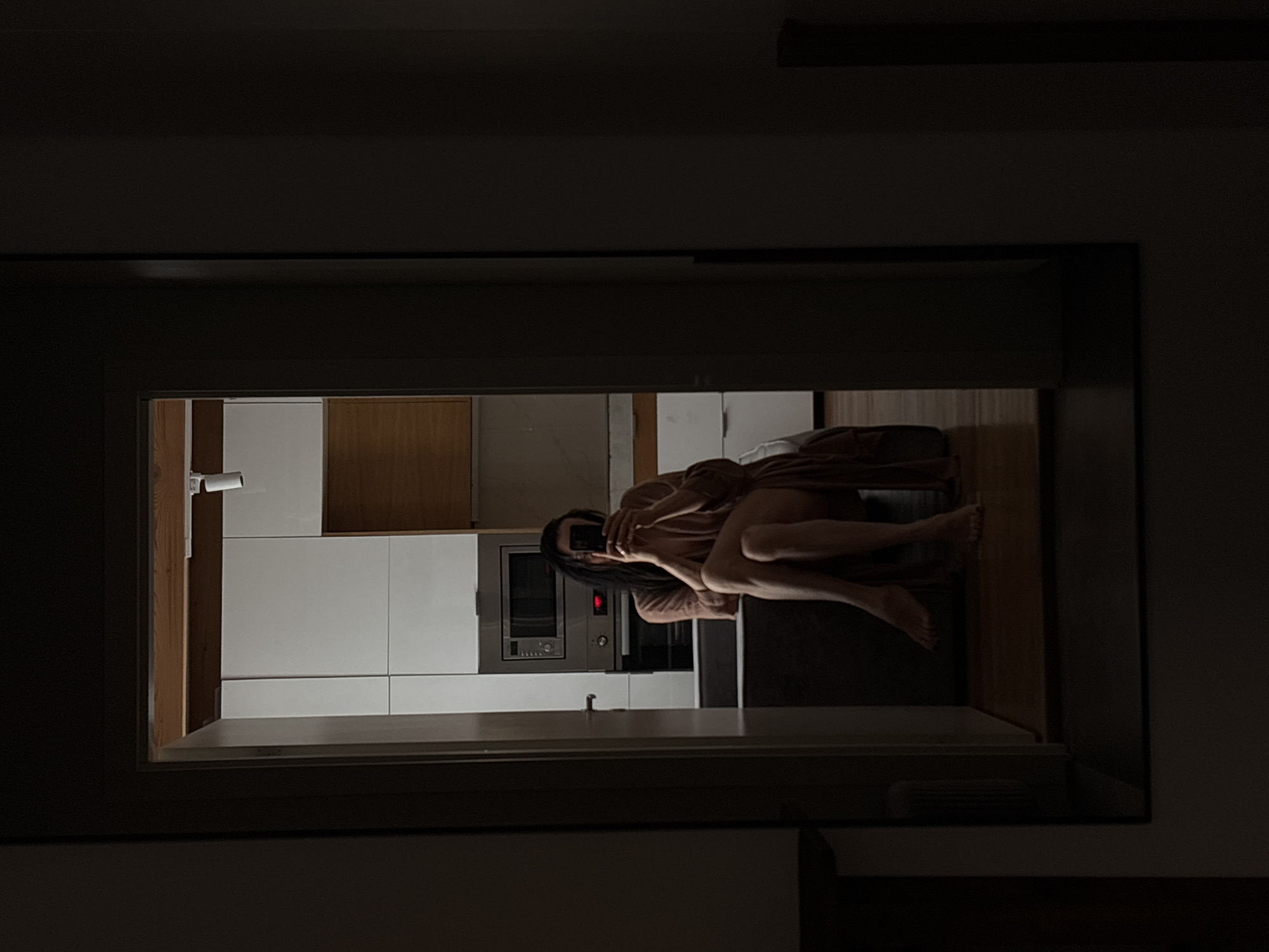 A dimly lit photograph shows a woman seated, facing the viewer, and illuminated primarily from a light source within an adjacent kitchen. The view of the woman and the kitchen are framed by a dark doorway. The woman has long, dark hair and is partially covered by a brown garment or blanket. Her legs are visible. The kitchen features white cabinetry, wooden accents, and a built-in microwave. The image appears to have been taken indoors at night. The location is Chaikovsky, Russia.