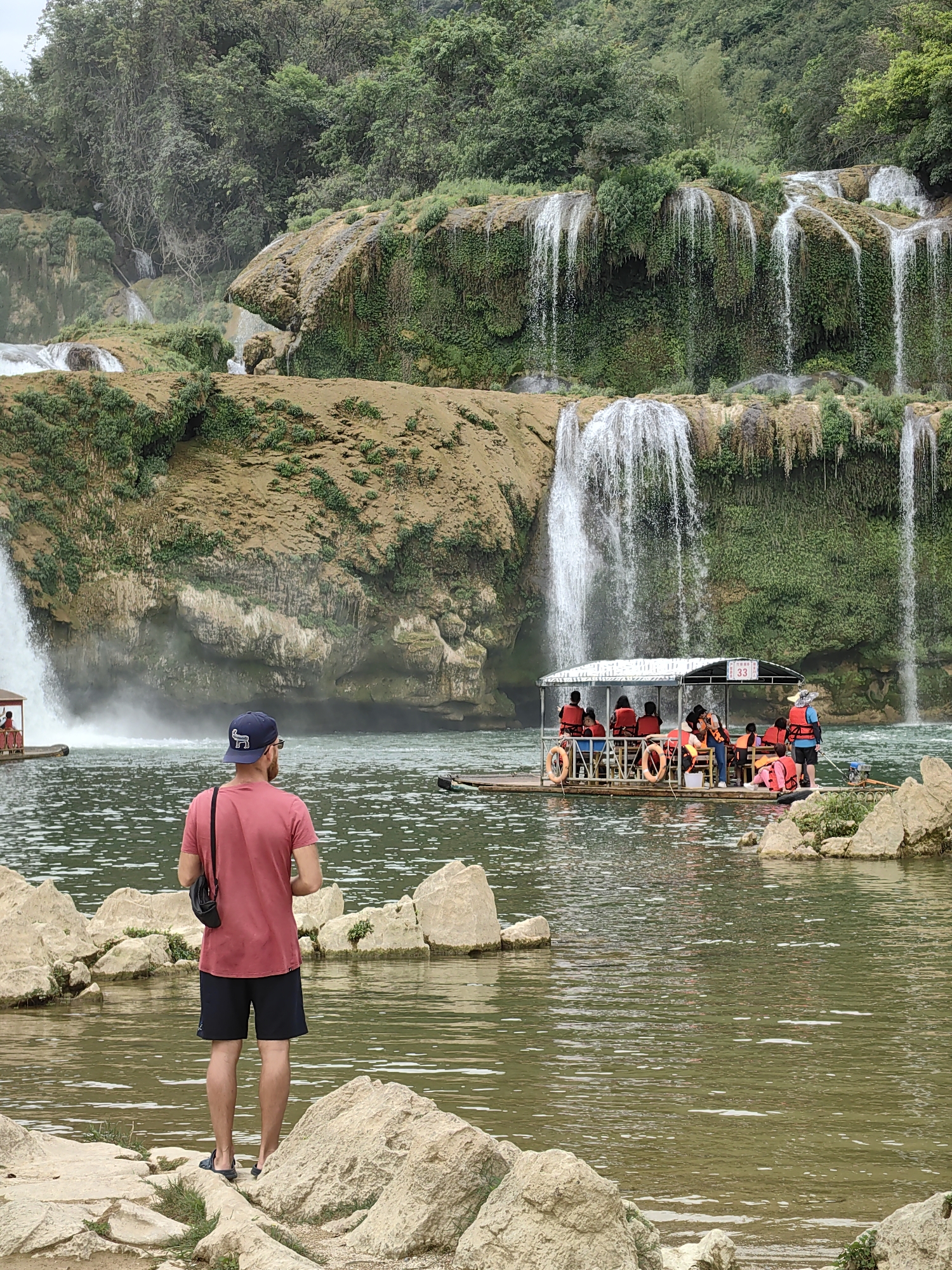 A full shot captures a man standing on a rocky shore facing a body of water with waterfalls in the background in Daxin County, China. The man is seen from the back, wearing a red shirt, black shorts, a blue baseball cap, and black sandals. He has a black bag slung over his shoulder. The body of water contains a raft carrying several people. The people on the raft are wearing orange life vests. The background features tiered waterfalls cascading over mossy rocks and lush green vegetation. The sky is overcast. The water appears murky green in color. The scene conveys a sense of natural beauty and tourism.
