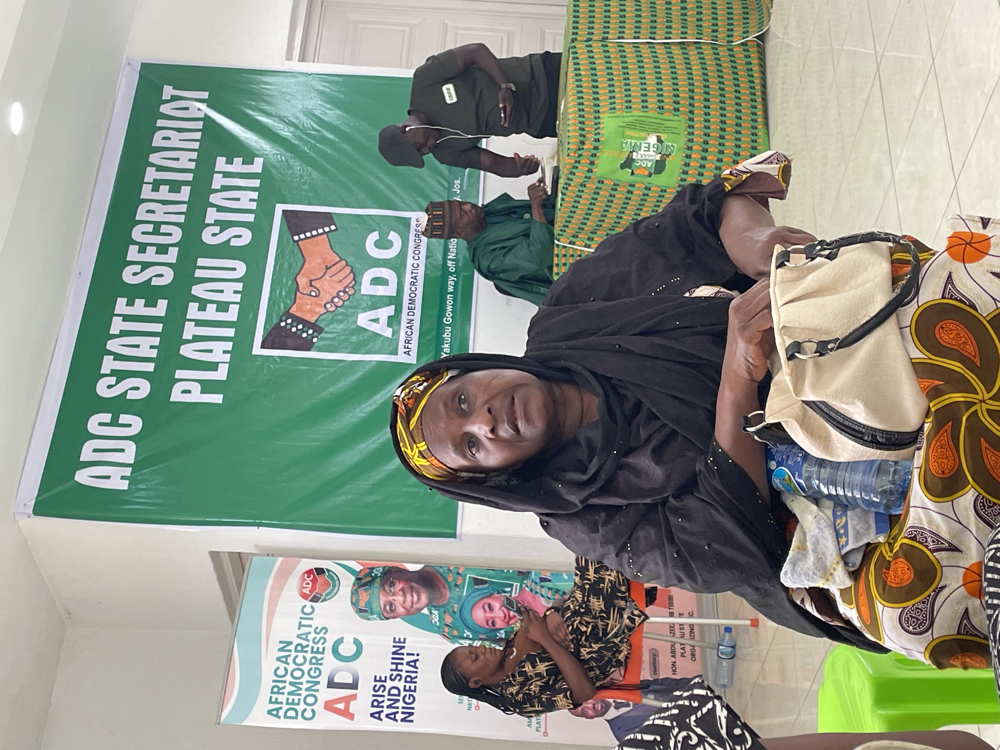 The image shows a gathering of people associated with the African Democratic Congress (ADC) in Jos, Nigeria. A woman in a black hijab sits prominently in the foreground. Banners with ADC logos and slogans like "Arise and Shine Nigeria!" are visible. One banner specifically identifies the location as "Yakubu Gowon way, off Natio J, Jos." Several other individuals are present, including men in traditional attire and a man in a green shirt with headphones. A woman is seated and engaging with her phone. Objects such as water bottles, bags, and plastic chairs are scattered around the scene.