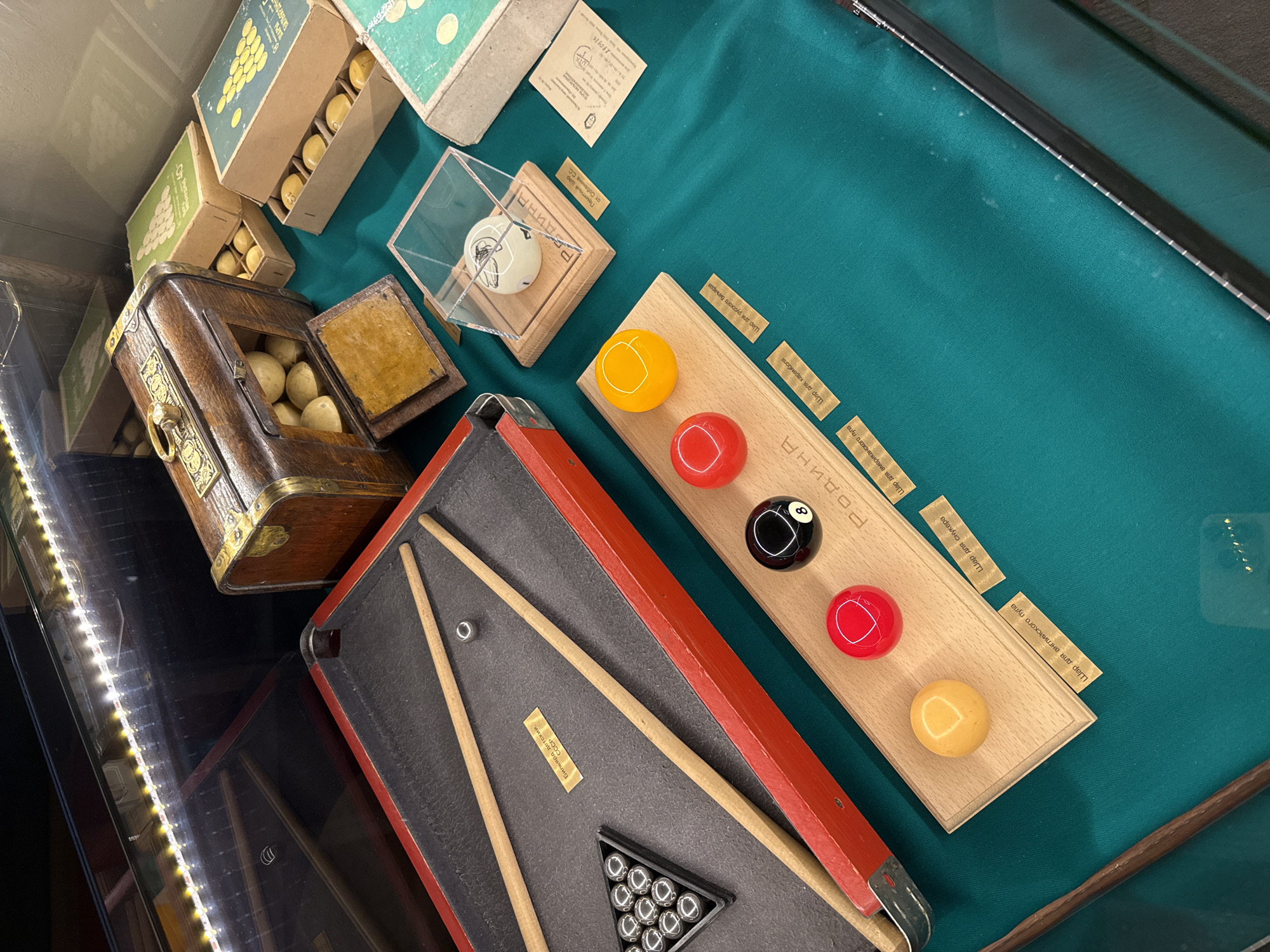 The image depicts a display of antique billiards equipment, likely in a museum or exhibition. The items are arranged on a green felt surface, reminiscent of a billiards table. In the foreground, a miniature red-rimmed billiards table with two cues and a rack of silver balls is visible. Next to it, a wooden board displays four billiard balls: a yellow one, two red ones, and a black eight ball. There are also several small labels with Cyrillic text associated with the balls. In the background, a vintage wooden chest filled with small, round, pale objects is open. Several green boxes containing billiard balls are also present, with some balls visible inside. A glass case encloses a white billiard ball with a drawing on it. A document is visible to the right of the glass case. The lighting suggests an indoor environment, possibly a museum display with overhead lights illuminating the scene. The overall impression is that of a historical collection of billiards memorabilia.