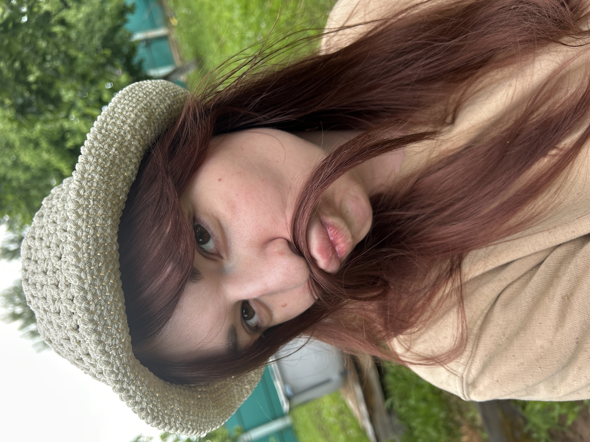 The image presents a close-up portrait of a woman outdoors. She has long, reddish-brown hair, some of which falls around her face and over her right eye. She is wearing a light, textured hat, possibly crocheted or knitted, in a neutral light beige or pale green shade. A light beige or tan colored garment, resembling a hoodie or sweatshirt, is visible on her shoulder and arm.
The woman's head is tilted, and she looks directly at the camera with her eyes open. Her lips are puckered in a "kissing face" or "duck face" expression, conveying a playful or casual mood.
The background is softly blurred but shows an outdoor setting with ample green foliage, indicative of trees and grass. Partially visible man-made structures in the background include a green fence or shed and another light-colored object. The lighting suggests natural daylight, without strong shadows. No text is visible in the image. The location context for the scene is Фоки, Russia.