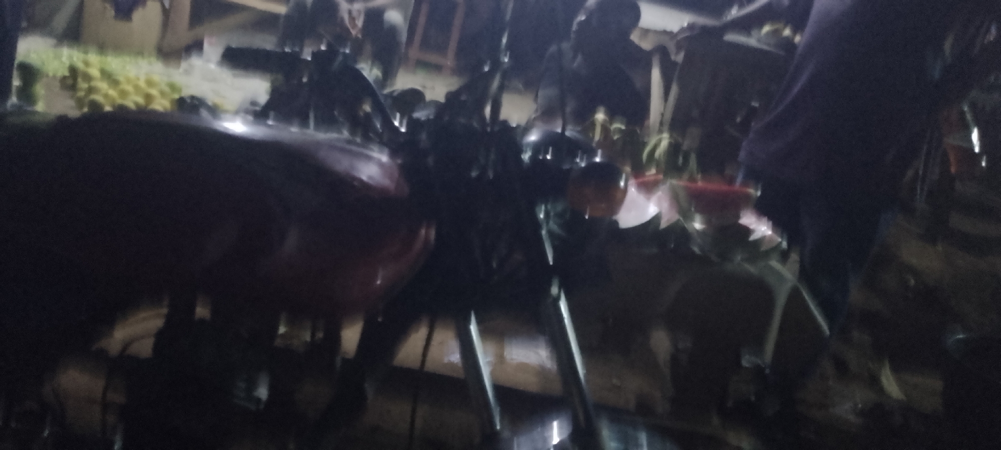 The image depicts a dimly lit outdoor scene, likely a market or street stall, in Jalingo, Nigeria. In the foreground, a red motorcycle is partially visible, its details obscured by motion blur and low light. The motorcycle appears to be parked or stationary, with its handlebars and front section clearly discernible. Behind the motorcycle, several people are present, their forms indistinct due to the low light and motion blur, suggesting activity and movement within the scene. The background reveals what appears to be goods for sale, possibly fruits, arranged on tables or stalls, also affected by the poor lighting conditions. The overall impression is of a bustling, albeit dimly lit, marketplace in Jalingo, Nigeria, with a prominent red motorcycle as a focal point.