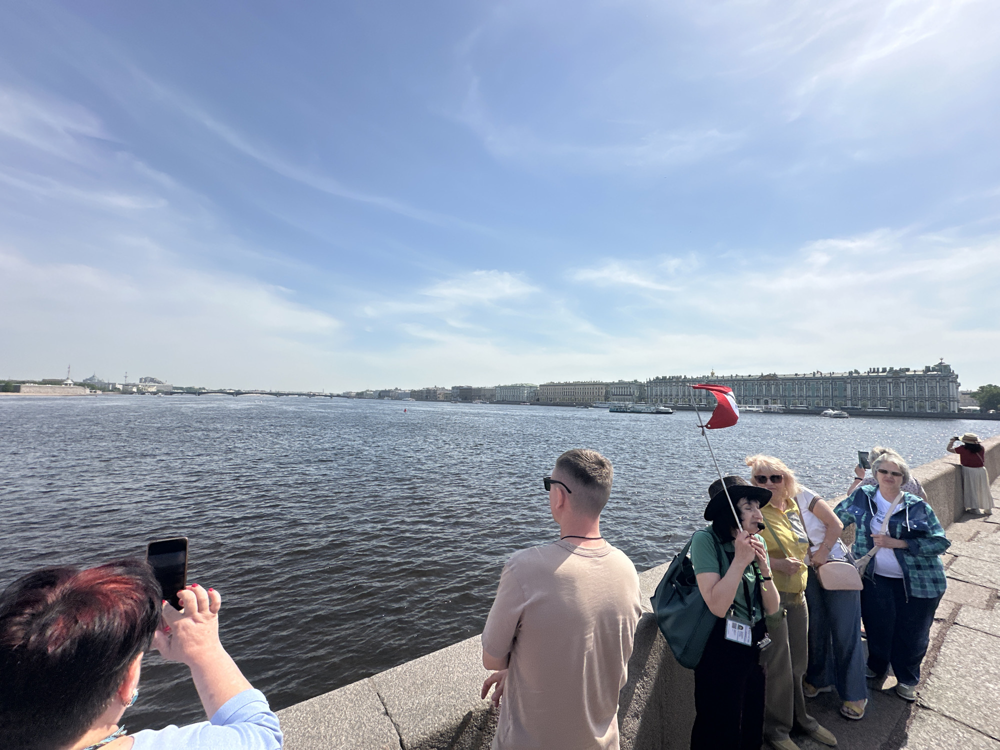 The scene captures a group of tourists on a bright day, possibly summer, in Saint Petersburg, Russia, observing the waterfront of the Neva River. The composition includes the vast expanse of the river, the cityscape across the water, and a cluster of people on what appears to be a stone embankment or bridge.
To the left of the frame, a woman is holding up a cell phone to take a picture, her focus on the river view. Beside her, a man with short-cropped hair and sunglasses stands with his back to the viewer, gazing at the cityscape. Further right, a group of women are gathered, one holding a small red and white flag, possibly as a tour guide. The women are dressed in casual clothing, including a plaid jacket, olive pants, and a black sunhat. The backdrop features historical buildings and boats on the river, quintessential of the city's landscape. The sky is a clear blue with streaks of wispy clouds. The general atmosphere is of tourism and sightseeing.