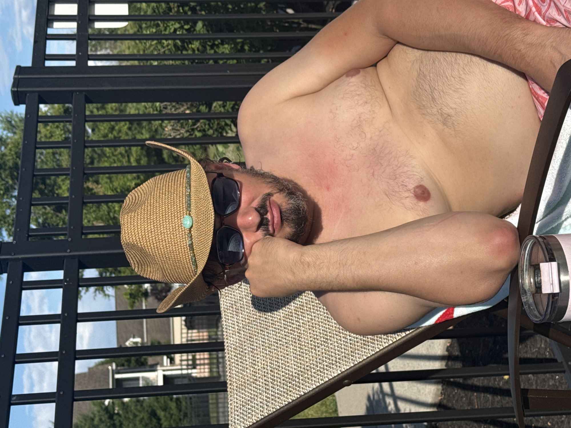 A Caucasian man with a beard and a tan cowboy hat with a turquoise embellishment is sitting in a lounge chair outdoors, leaning on his hand and wearing sunglasses. He is shirtless, exposing his chest and arms, and appears to have a sunburn on his chest. He is wearing pink patterned shorts. A metal fence with vertical slats is visible behind him, with trees and houses in the background. A tumbler cup is in the foreground on the chair. The setting is likely a residential backyard or poolside area in Allen Township, United States of America.