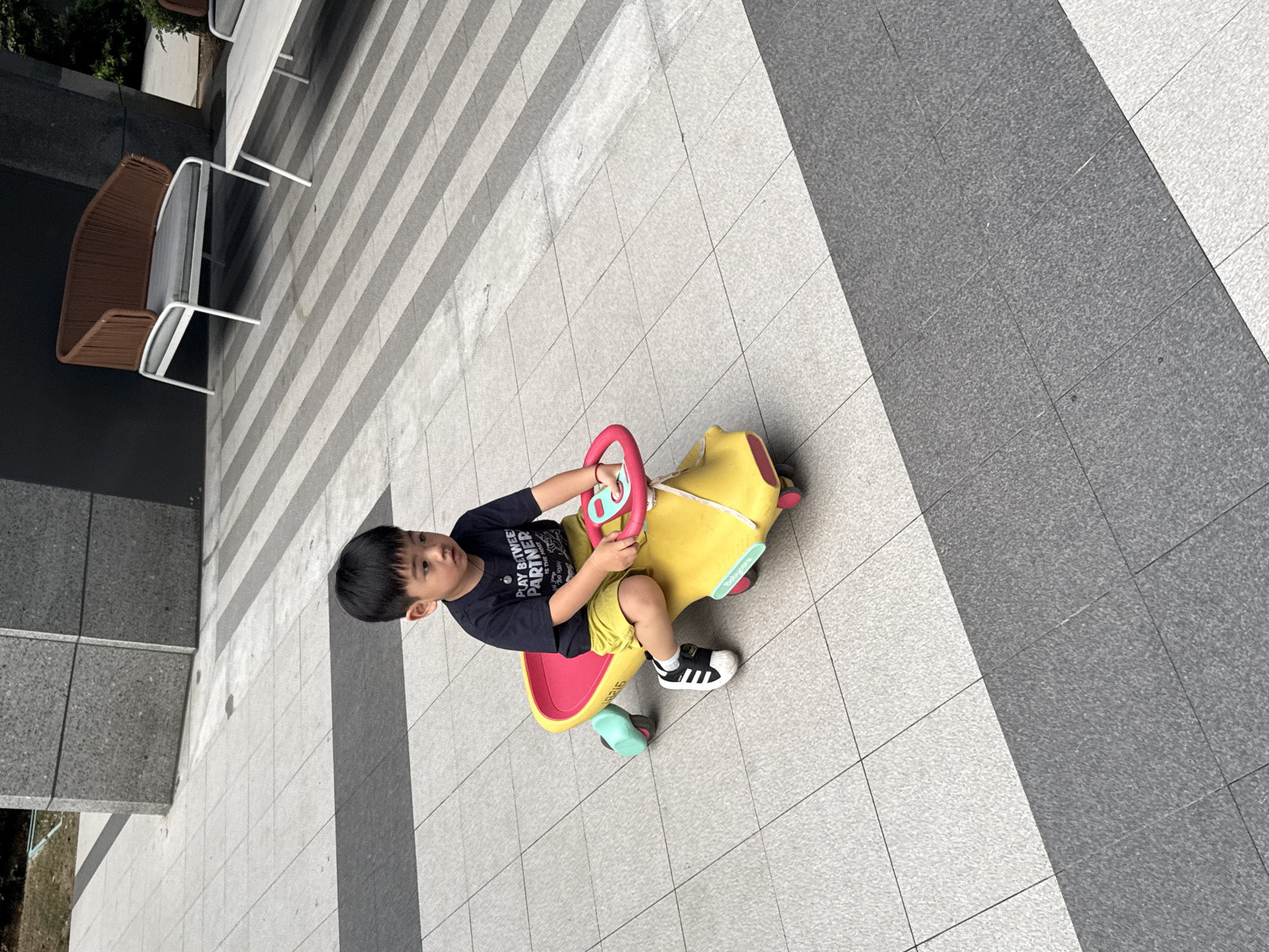 An Asian child, appearing to be around 3-4 years old, sits on a yellow ride-on toy car with a red steering wheel. The child is wearing a black t-shirt with white text that reads "PLAY BETWEEN PARTNER IS THE HOST" and yellow shorts. They are also wearing black and white Adidas sneakers. The child is looking down and slightly to the right, with their hands on the steering wheel. The ride-on car has two teal-colored wheels visible. The child is on a tiled outdoor patio with a pattern of alternating light and dark grey square tiles, interspersed with dark grey rectangular strips. In the background, there are modern chairs and tables, and a building with a grey stone facade. The scene appears to be in Yiwu, China, likely an outdoor seating area or a public space within a commercial or residential complex.