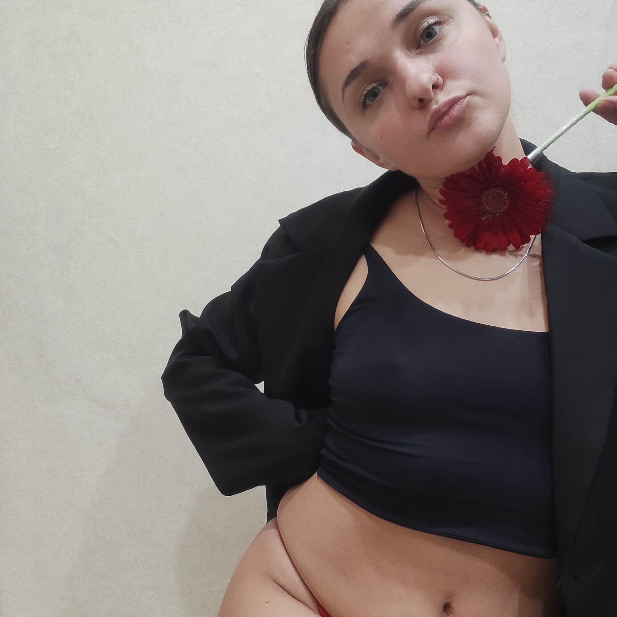 A young woman is posing in front of a plain wall. She is wearing a black crop top, a black blazer, and red underwear. She has a silver necklace and a silver nose ring. She is holding a red gerbera daisy near her neck. The location is not specified in the image.