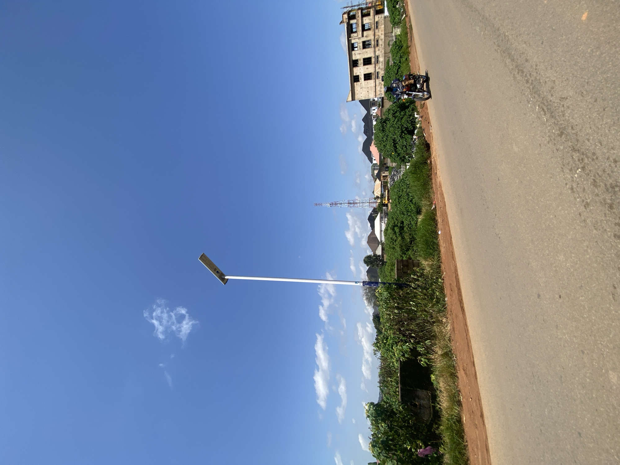The image captures a street scene in Gura Top, Nigeria, under a clear blue sky dotted with scattered white clouds. A solar-powered street lamp stands tall on the right side of the paved road. A person on a motorcycle is traveling along the road, moving from left to right, with a building under construction visible in the background. The area is characterized by lush green vegetation, and other buildings and a telecommunications tower can be seen in the distance. The road is asphalt, with a dirt shoulder on the right side.