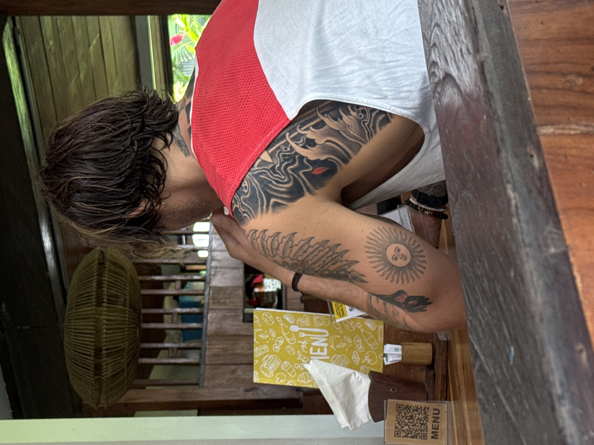 The image, rotated 90 degrees clockwise, depicts a person seated at a wooden table within an open-air restaurant or cafe, likely in North Kuta, Indonesia. The individual, viewed from behind and the side, has dark, wavy hair and is wearing a white sleeveless top with a prominent red mesh panel on the back.
The person is extensively tattooed. On their upper back and shoulder, an intricate black and white design features swirling patterns and a small red element, possibly depicting lips. The upper arm showcases a large tattoo of a sun with a face, reminiscent of the "Sun of May," surrounded by rays. Below this, another tattoo displays a stylized feather or leaf pattern, and further down the arm, a black and white flower resembling a calla lily or tulip. A beaded bracelet is visible on the person's wrist.
On the wooden table, a bright yellow menu is prominently displayed, adorned with white line-art icons of various food and drink items (e.g., burgers, beer, coffee). The word "MENU" is clearly visible on it, with partial text "SPECIAL... 7AM-11" also discernible. A white paper napkin, folded into a triangle, rests beside the menu. Additionally, a small wooden stand with a black QR code and "MENU" printed vertically is on the table.
The background shows wooden architectural elements, including vertical slats and beams, and lush green foliage with hints of pink flowers, indicating an outdoor or semi-outdoor setting. To the left, a woven, oval-shaped object, possibly a light fixture or fan, is visible. The scene suggests the person is engaged in a typical dining activity, such as reviewing the menu or waiting to order.