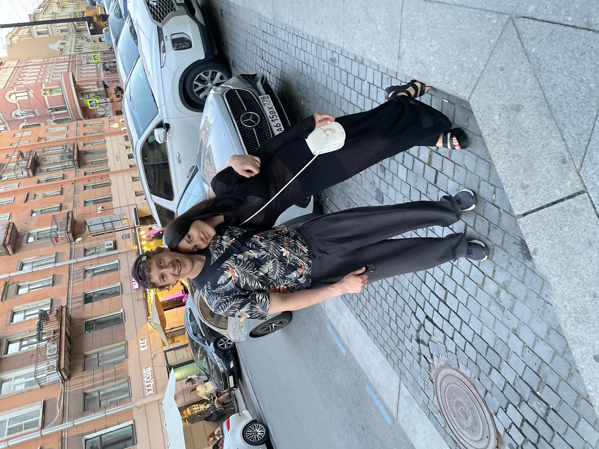 The scene depicts an urban street in Saint Petersburg, Russia, with two individuals posing for a photograph in the foreground. The man, of European descent, stands on the right, his right arm around the woman's shoulders. He wears a dark, backward-facing baseball cap, a dark short-sleeved shirt with a tropical floral pattern, dark loose-fitting trousers, and dark sneakers, with a dark strap crossing his chest. He smiles broadly at the camera. The woman, also of European descent, leans her head against the man's right shoulder, her left arm wrapped around his waist. She wears a long, sheer black dress or tunic over dark undergarments, and black platform sandals. She holds a small, off-white crossbody bag with gold-toned hardware, her fingernails painted light pink. She smiles subtly.
Behind the couple, a silver Mercedes-Benz sedan is parked, displaying the Russian license plate "A615BX 778," which indicates the Saint Petersburg region (778). Behind the Mercedes is a white Hyundai Palisade SUV. The street features a grey cobblestone sidewalk, which is uneven in places, and a circular metal manhole cover is visible on the left. A blue painted line marks the edge of the asphalt road.
The background consists of multi-story, historic European-style buildings with facades predominantly in shades of peach, orange, and light pink. These buildings feature numerous windows, balconies with metal railings, and visible air conditioning units. Storefront signs are visible, including "ХАЛКОН" (HALKON), "БАР" (BAR), and partially visible "СУШИ" (SUSHI). Further down the street, outdoor cafe seating with white umbrellas and several indistinct figures are present. The lighting suggests late afternoon or early evening.