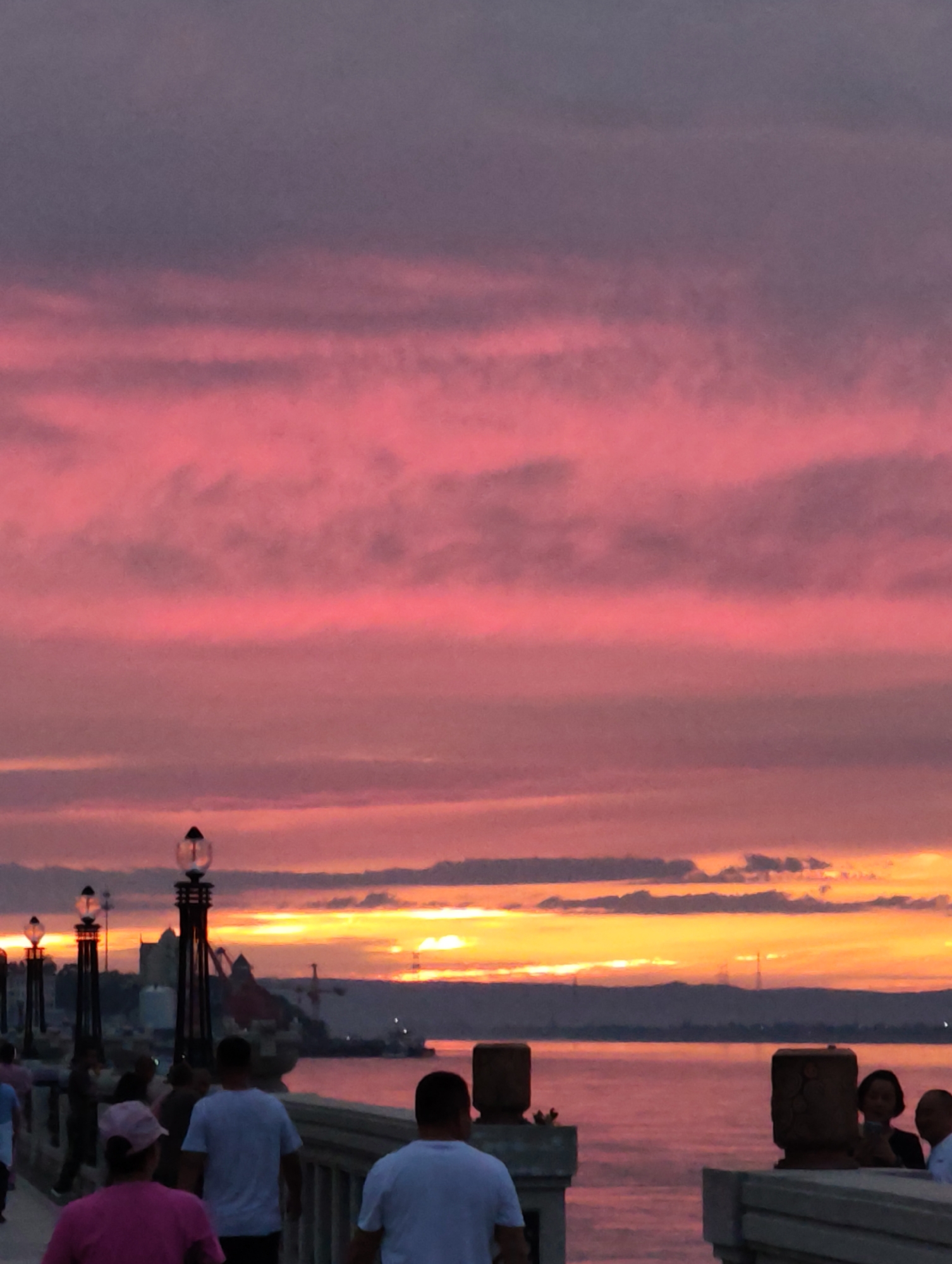 A sunset paints the sky with vibrant hues of pink and orange over the Aihui district of China. The horizon is marked by a distant cityscape, silhouetted against the colorful sky, with a body of water in the foreground. A pedestrian bridge spans across the water, with several people walking along it. The bridge is lined with decorative lamps, casting a warm glow on the scene. The people on the bridge are casually dressed and appear to be enjoying the view of the sunset. The overall atmosphere is serene and picturesque.
