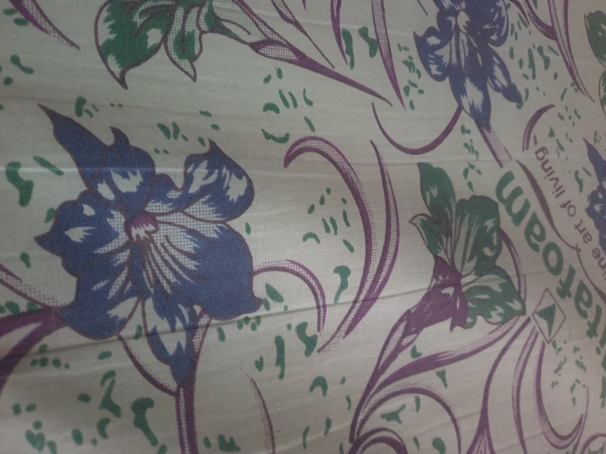The image shows a close-up, abstract view of a patterned fabric, likely a bedsheet or curtain, with stylized floral motifs in shades of blue, green, and purple against a pale background. The pattern features large, open flowers with pointed petals and swirling vine-like elements. Scattered green dots and dashes add texture to the design. In the lower portion of the image, text is partially visible, reading "vitfoam" in green lettering, followed by "fine art of living" in smaller white lettering. The fabric appears slightly creased, suggesting it might be draped or folded. The lighting is diffuse, and the focus is on the intricate details of the pattern. There is no discernible indication of the location in Jalingo, Nigeria, as the image is purely an artistic representation on fabric.