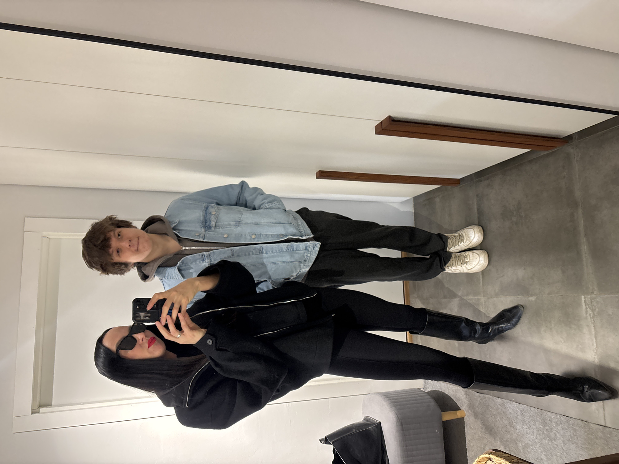 A selfie, likely taken in a store fitting room. Two individuals are present: one wearing a light-wash denim jacket with a grey hoodie underneath, dark trousers, and white sneakers; the other, assumedly the photographer, is wearing a black ensemble featuring a jacket, leggings, and knee-high black boots. The photographer is holding a smartphone and wearing black sunglasses and red lipstick.
The background is a beige wall and a gray floor.
The location is Chaikovsky, Russia.