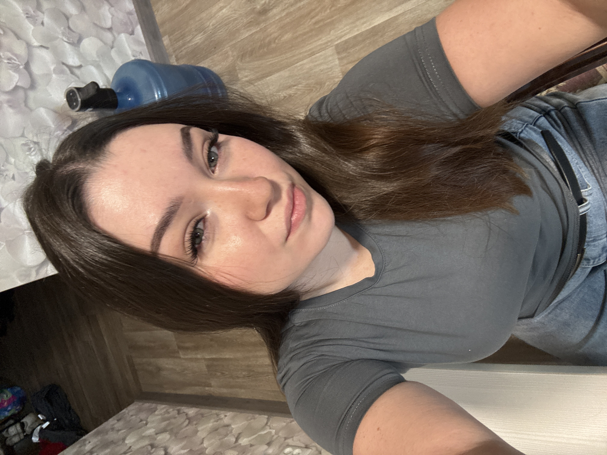 A young woman with long, dark brown hair is taking a selfie indoors. She is wearing a grey t-shirt and blue jeans. The background shows a wooden floor, a wall with floral wallpaper, and a large blue water cooler with a black dispenser. To the left of the image, some bags and clothing are visible on the floor. The lighting suggests it is daytime. The location appears to be a domestic setting in the city of Zarechnaya (Gamovskoe S/P), Russia.