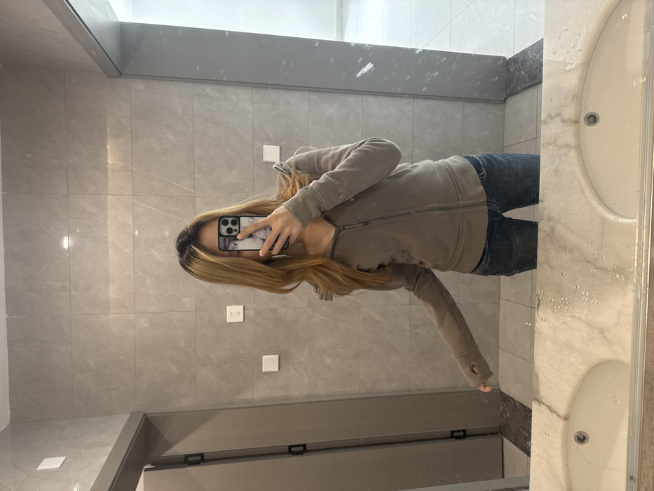 The scene depicts a single individual taking a mirror selfie inside what appears to be a public or private restroom in Nan'an District, China. The individual, a person with long, wavy, golden-blonde hair with dark roots, is partially obscured by a smartphone held in their right hand, positioned to capture the reflection. The smartphone has a dark frame and a light-colored, patterned case. The person is wearing a light brown/taupe zip-up hoodie with a hood and thumbholes in the sleeves, paired with dark blue denim jeans.
The environment features light grey, rectangular wall tiles with a subtle marble-like pattern. To the left, a medium brown-grey door with black hinges and a handle is visible. On the tiled wall, two white electrical outlets (one with two vertical slots, another with two vertical slots and a round ground hole) and two white light switches are mounted. The foreground is dominated by a large mirror reflecting the scene, beneath which a light-colored marble-patterned vanity counter is present. Two white oval sinks are partially visible below the mirror's edge, and water droplets are discernible on the mirror surface above the left sink. The floor appears to be covered in light-colored tiles, with a darker baseboard or border strip visible in the reflection. The room is brightly lit.