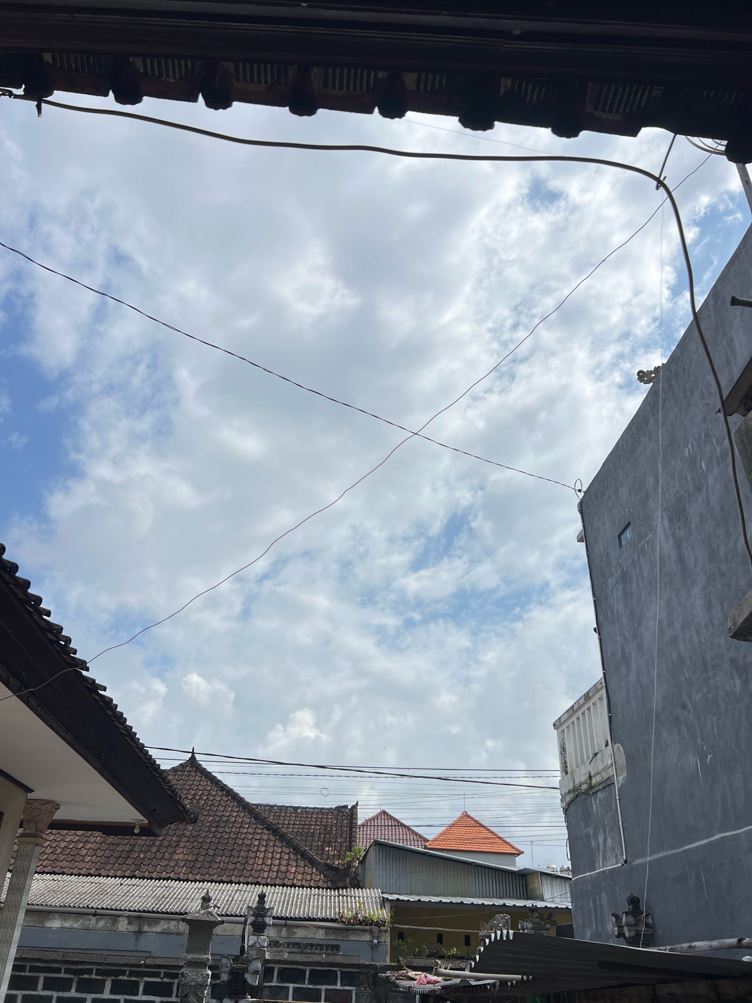 This is a photograph taken from a low angle looking up at the sky, with a foreground dominated by architectural elements. The image is characterized by a cloudy sky, with patches of blue visible. Two thin wires cross each other diagonally in the sky, creating a subtle X shape.
On the left side of the frame, a portion of a roof with dark brown tiles is visible, extending from the top edge of the image downwards. Beneath this roof, a glimpse of a white structure with decorative stone pillars suggests a building or a wall. Further in the background, a series of tiered roofs with reddish-brown tiles are prominent, characteristic of Indonesian architecture. A grey wall with a small, rectangular window is visible on the right side of the frame, also contributing to the urban residential feel. The overall impression is that of a residential area in an urban setting.
The presence of these architectural styles, particularly the tiered roofs and decorative pillars, strongly suggests the location is Denpasar, Indonesia. The style of the roofs and the general urban density are consistent with residential neighborhoods in Denpasar. The image captures a moment of an overcast day in what appears to be a typical residential district.