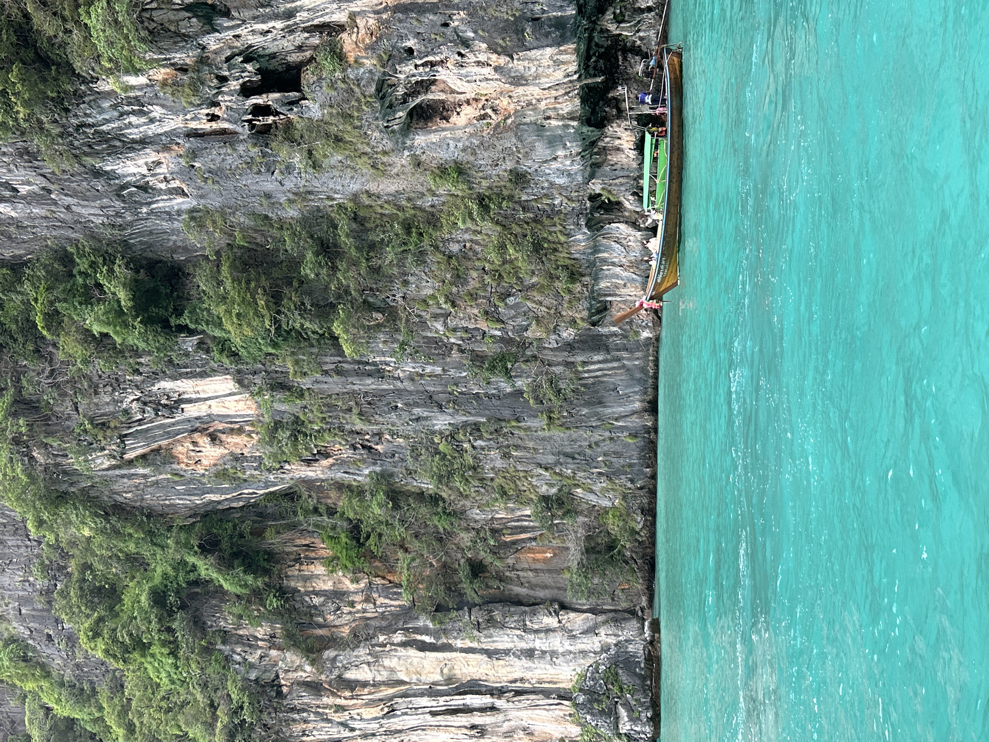 This image depicts a scenic view of towering limestone cliffs covered in lush green vegetation, bordering a vibrant turquoise body of water. A traditional Thai longtail boat, with a wooden hull and a green canopy, is seen navigating the clear water near the base of the cliffs. Several people are visible on the boat, suggesting it is in use for transport or a tour. The overall environment is characteristic of the coastal regions near Ao Nang, Thailand, known for its dramatic karst landscapes and stunning tropical waters. The lighting suggests it is daytime, with a soft, overcast sky, as indicated by the absence of harsh shadows.