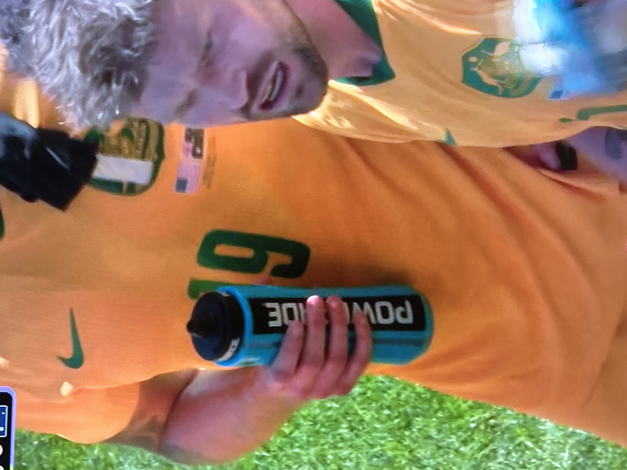 In a medium shot, a person wearing an orange soccer jersey with a green Nike logo and the number 19 is holding a blue Powerade water bottle. The bottle is positioned in front of the person's torso. To the right, another person in a yellow soccer jersey with a green collar is partially visible, facing towards the right side of the frame. The background shows green grass. The scene appears to be from a sporting event.